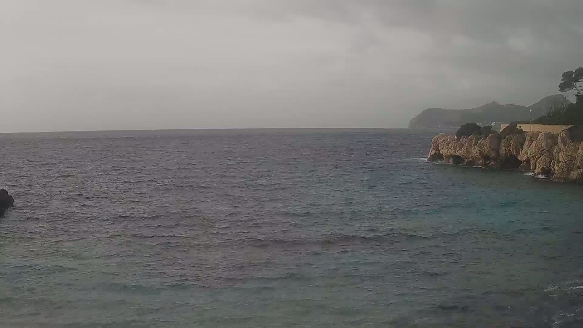 A rocky cliffside meets a calm, gray sea under an overcast sky.