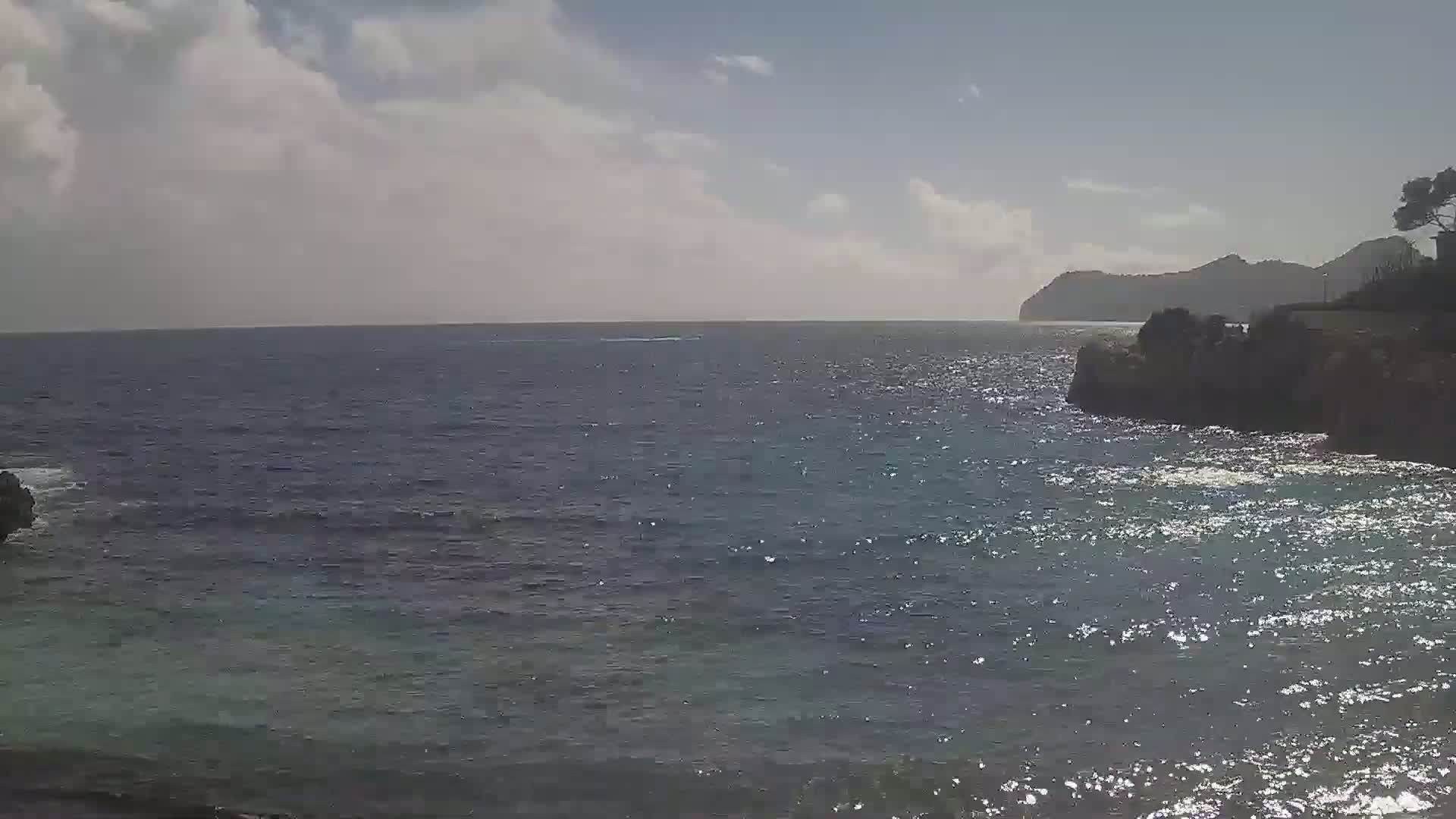 A partly cloudy day reveals a rocky coastline with dark blue, somewhat choppy water extending to a distant mountain range.