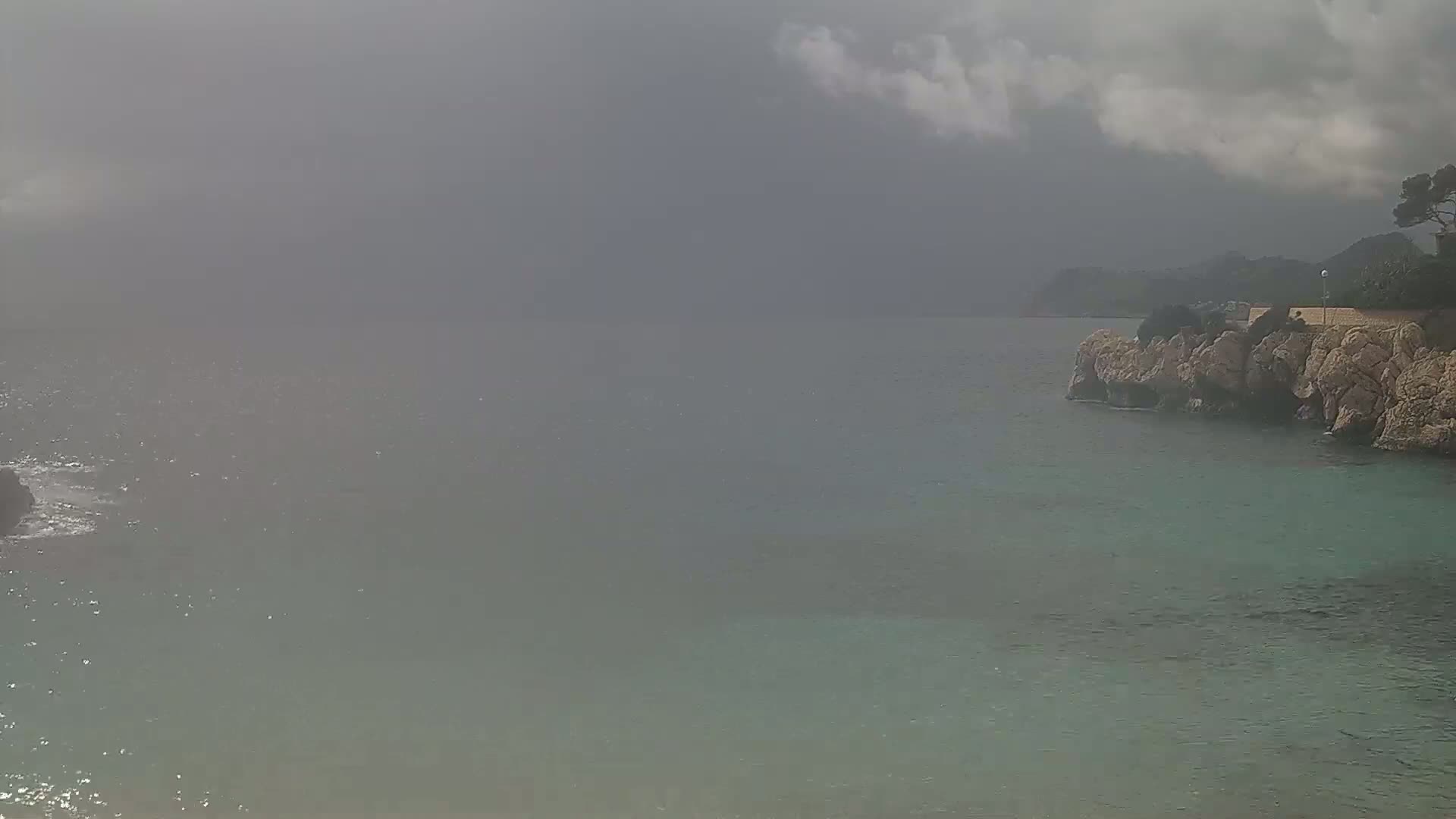 A rocky coast meets a calm, hazy sea under a mostly cloudy sky.