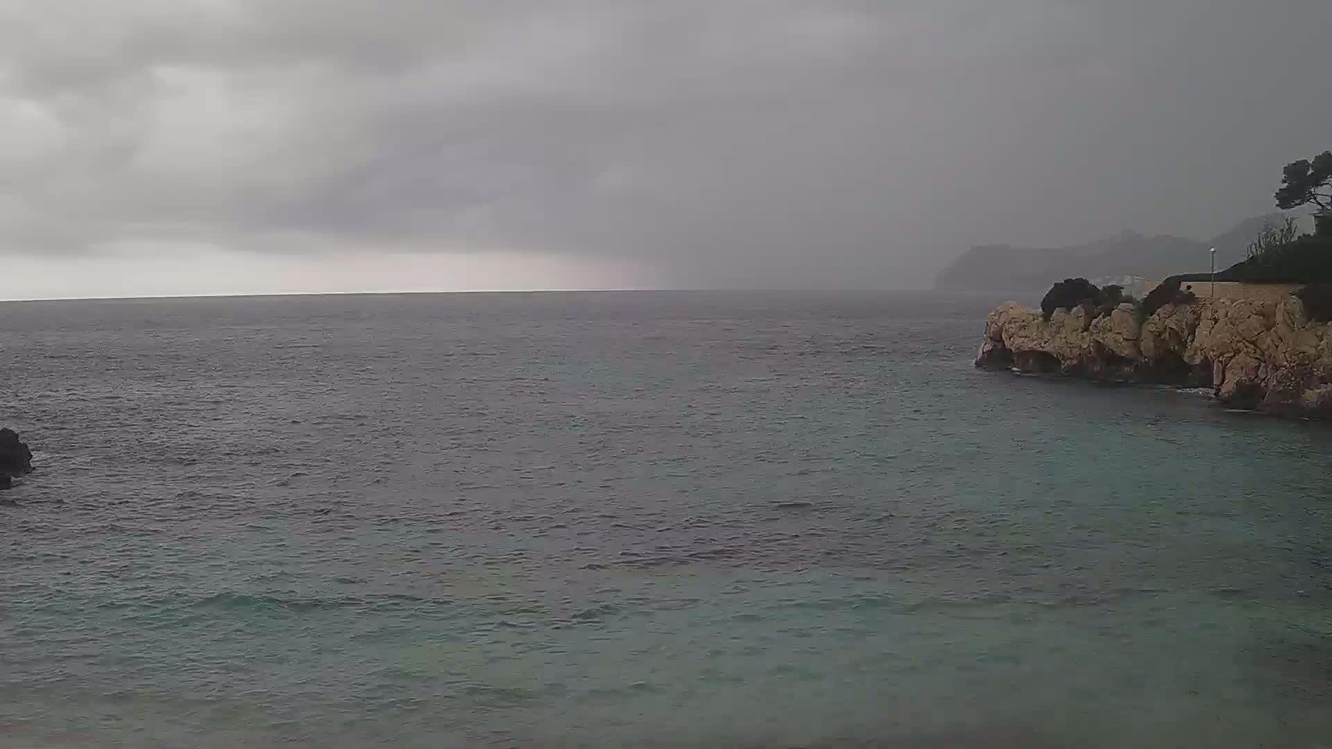 A dark, overcast sky hangs over a calm sea meeting a rocky coastline under a rainy sky.