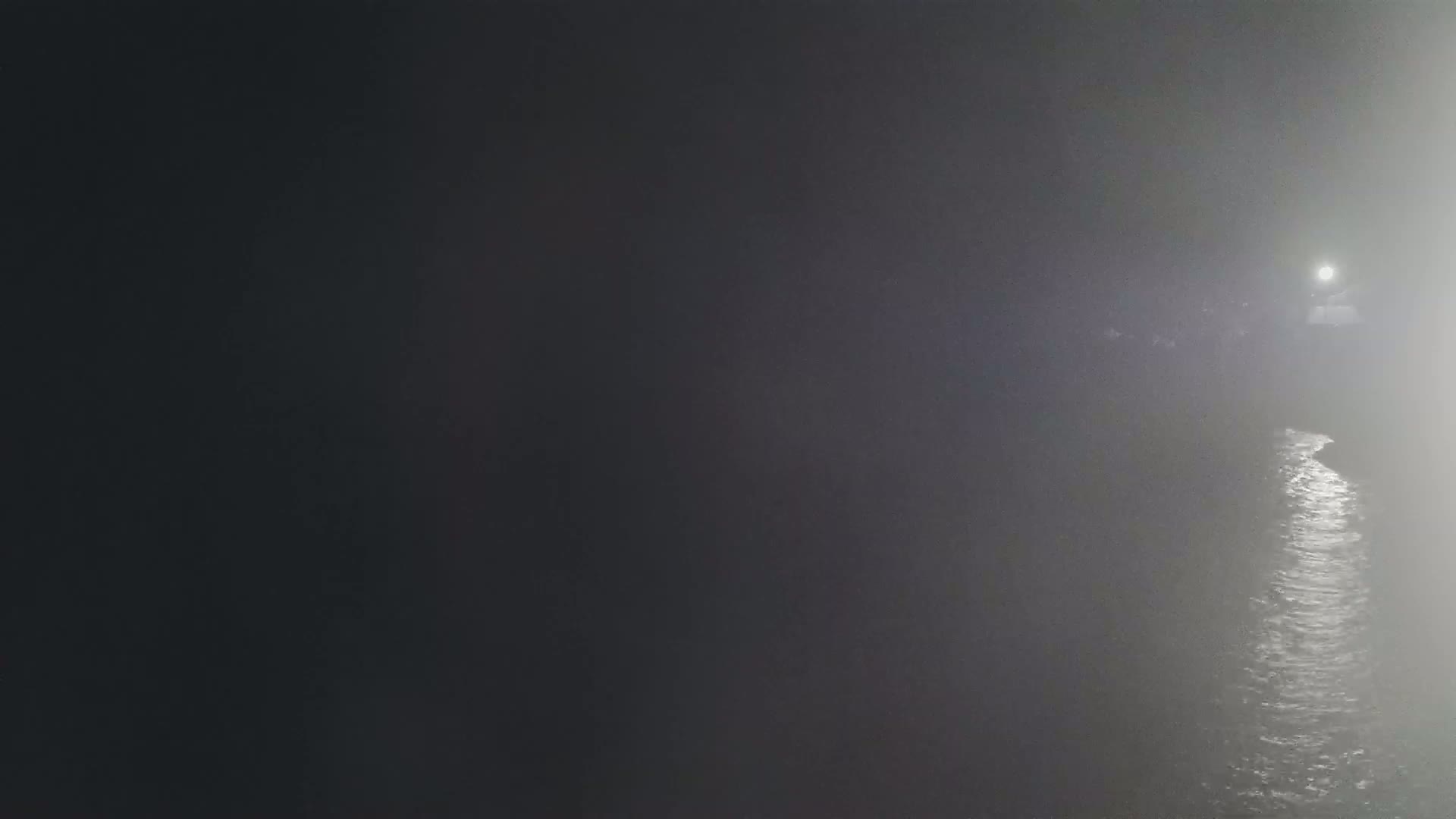 A dimly lit body of water is visible through a dense fog at night, with a single light source in the distance.