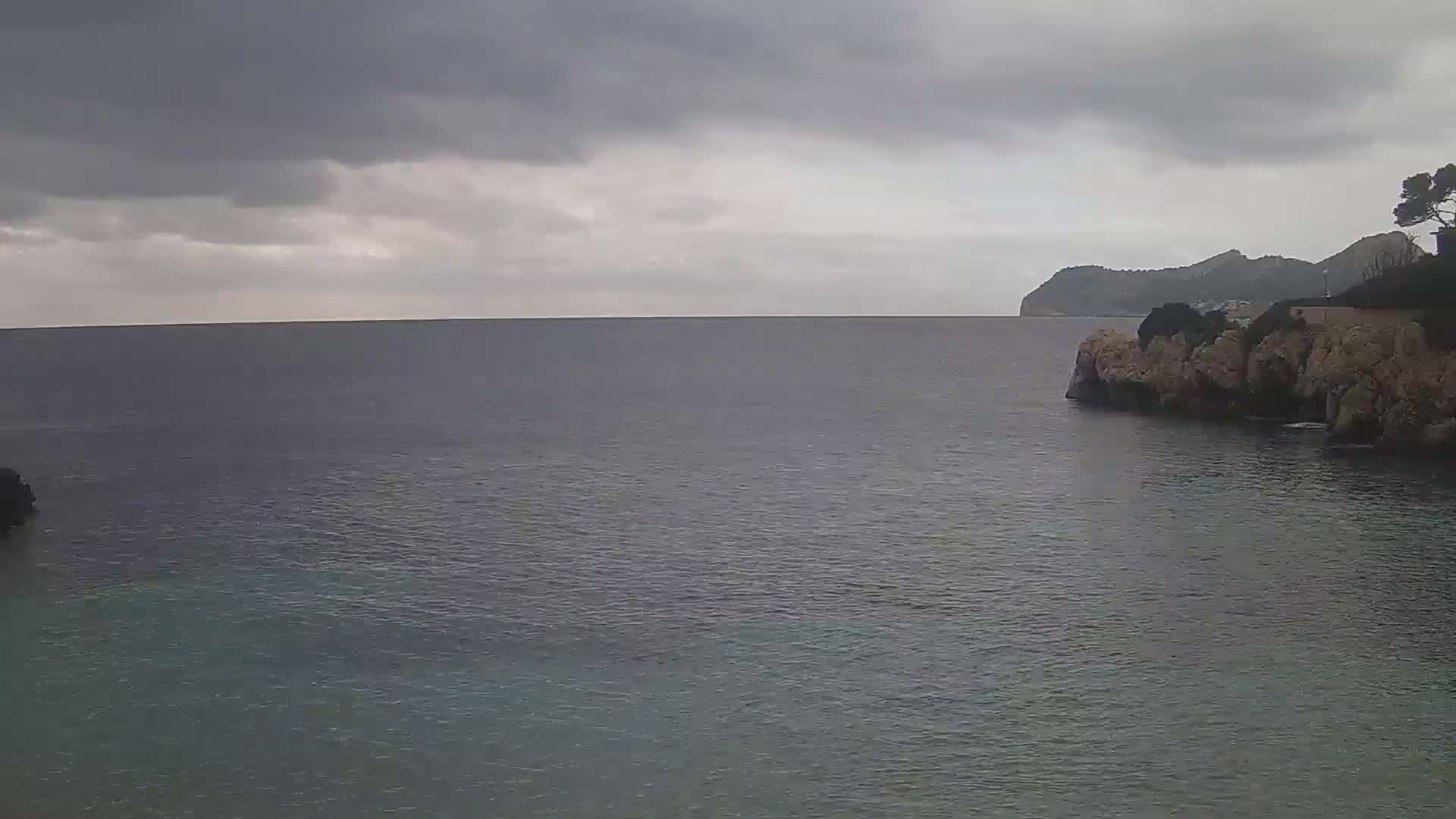 A calm sea under an overcast sky is partially enclosed by rocky shores, with a distant mountain visible in the background.