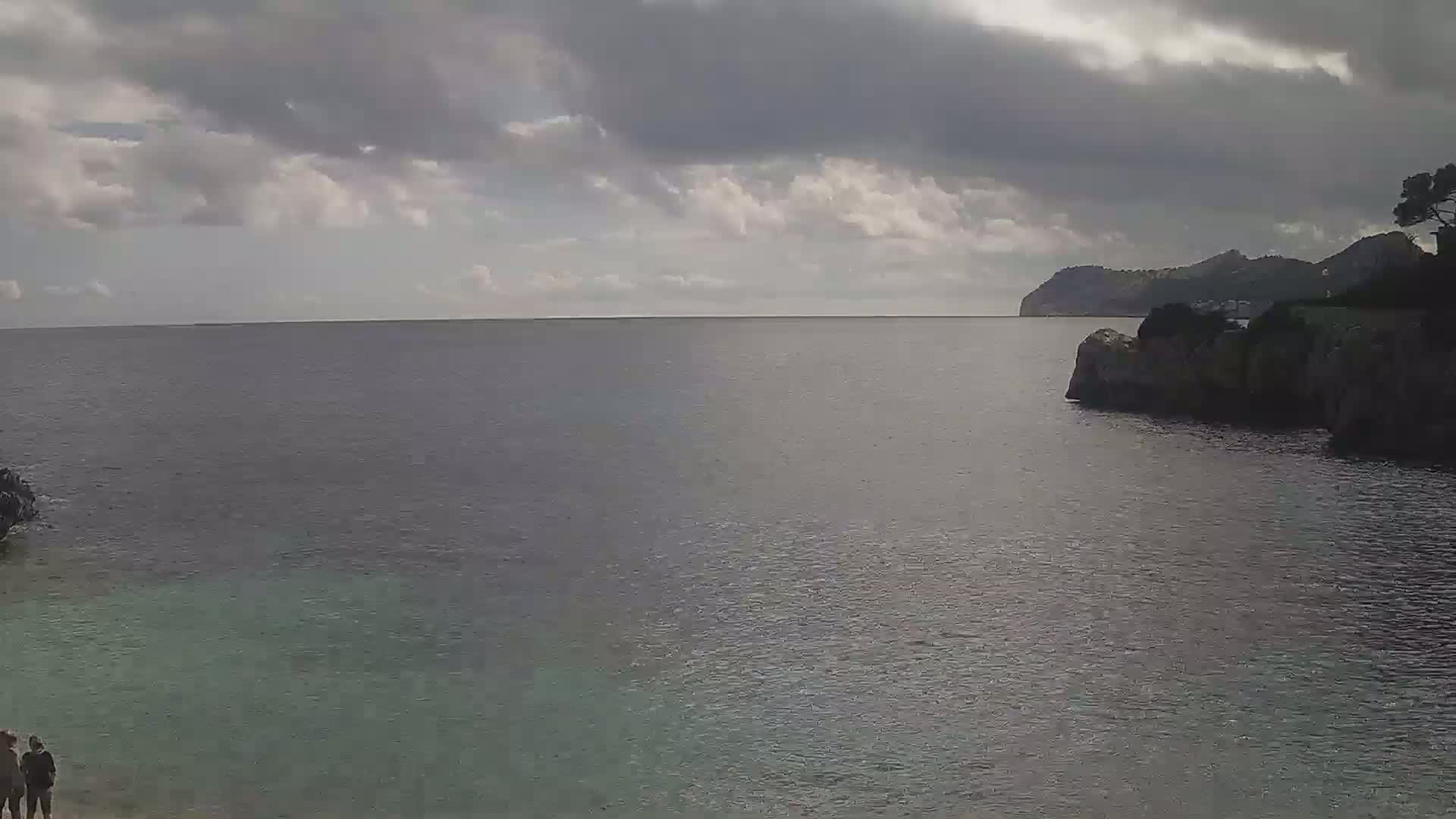 A calm sea under a cloudy sky is seen from a rocky shore, with a distant landmass visible and two people standing on the shore in the bottom left corner.