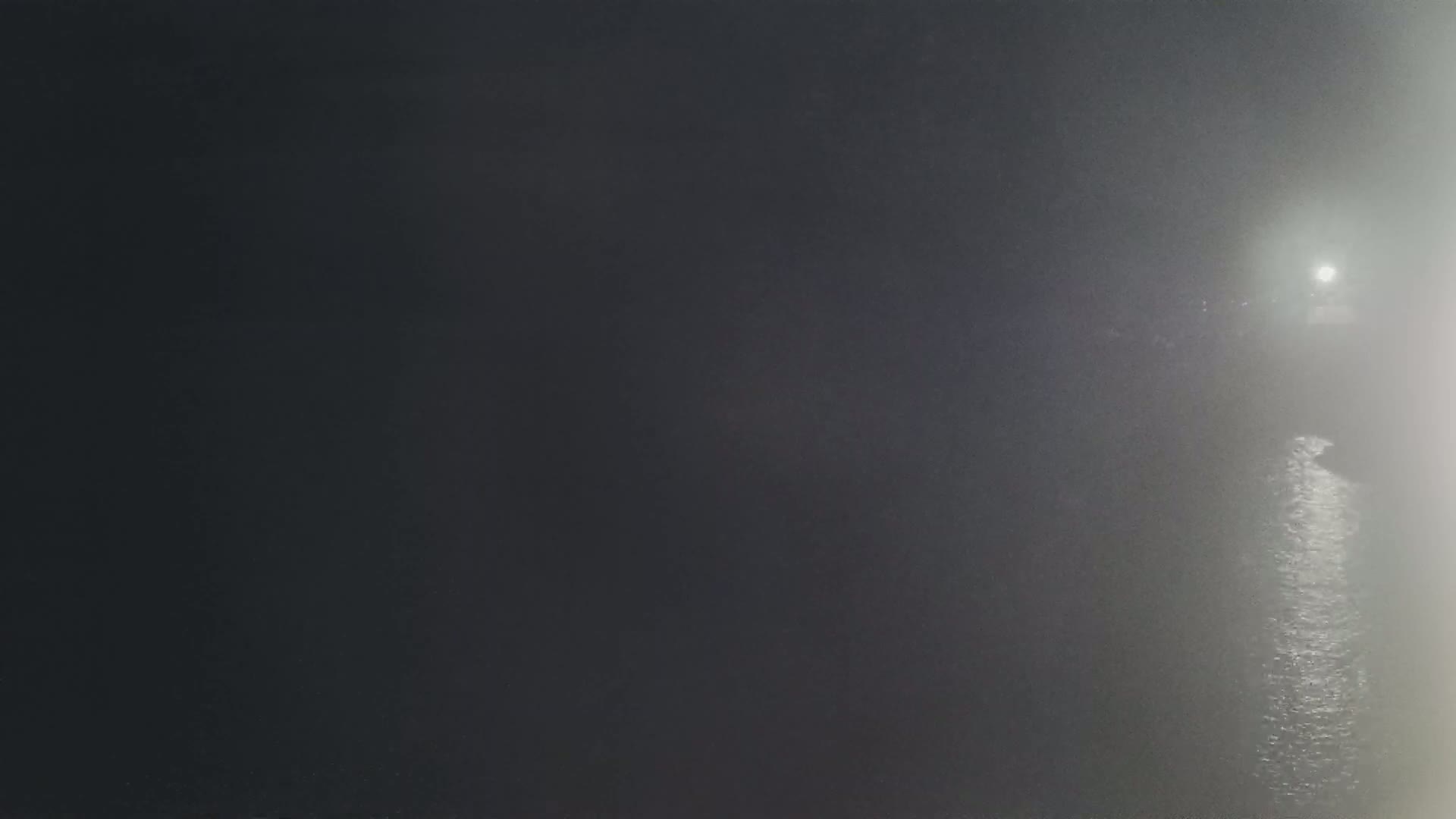 A dark, foggy night scene shows a body of water reflecting a distant light source.