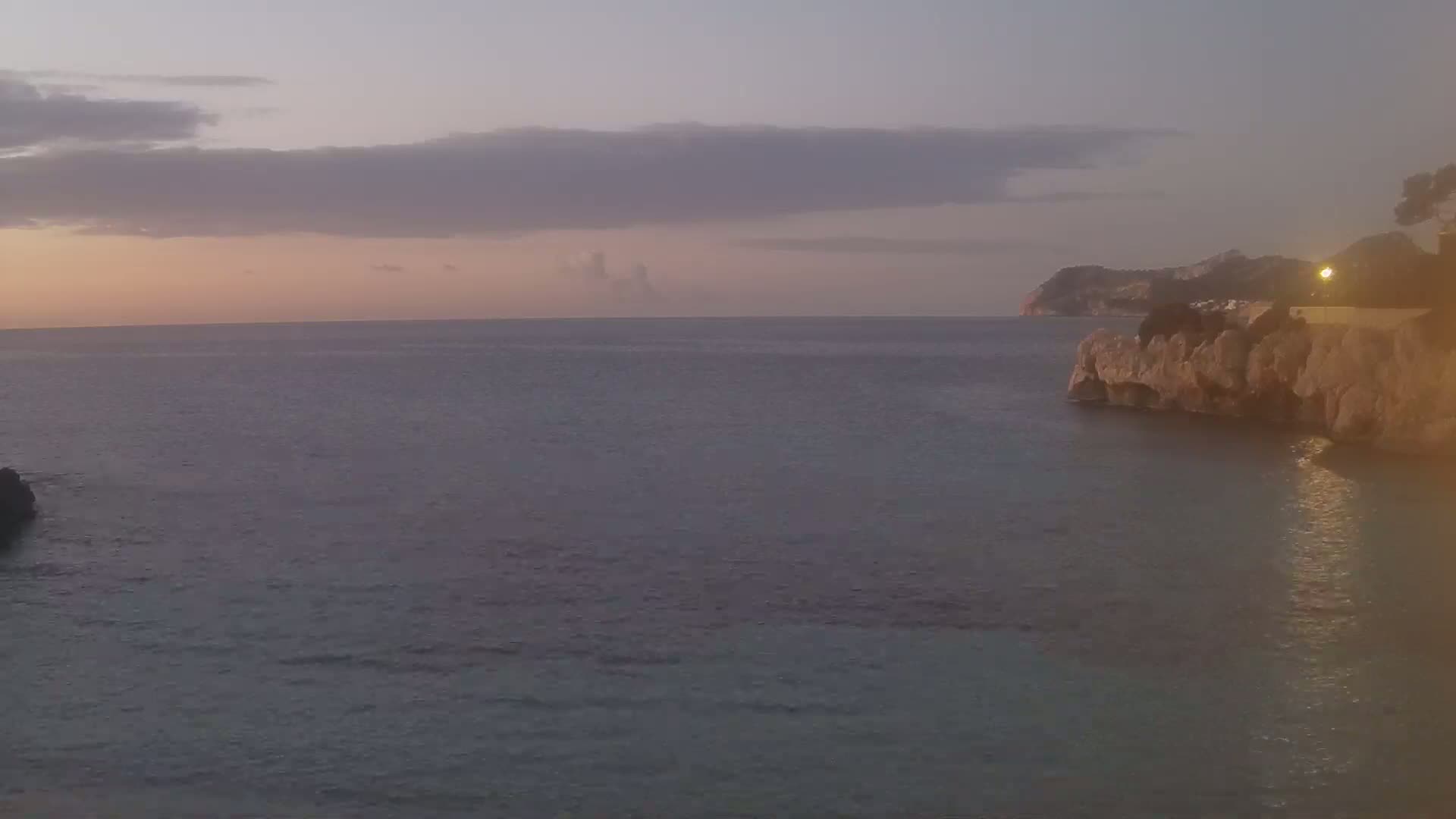 A calm sea at sunset is viewed from a rocky shoreline under a partly cloudy sky.