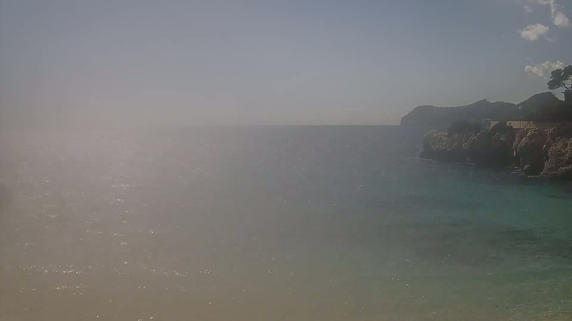 A hazy view of a calm sea meets a rocky coastline with a hill in the distance under a mostly clear sky.