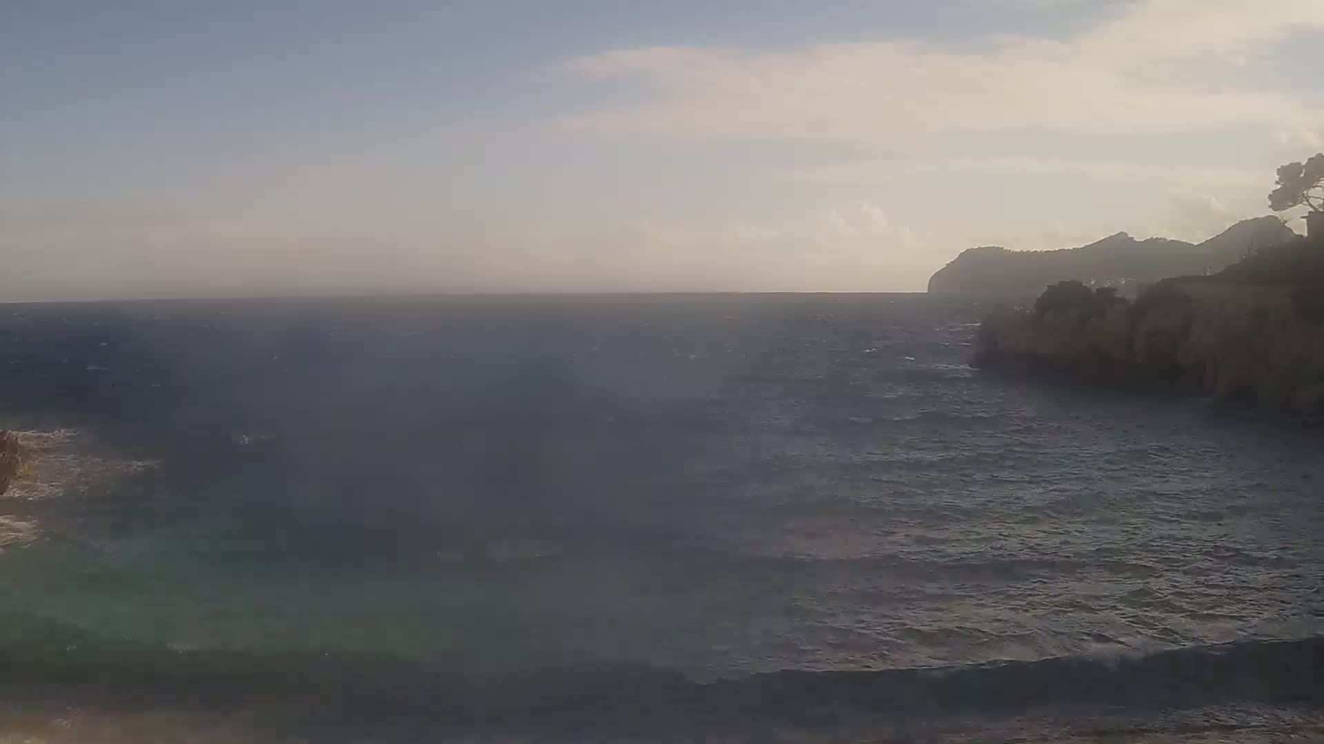 A slightly hazy, windy day shows a rocky coastline and dark blue, wavy ocean extending to a distant, silhouetted mountain.