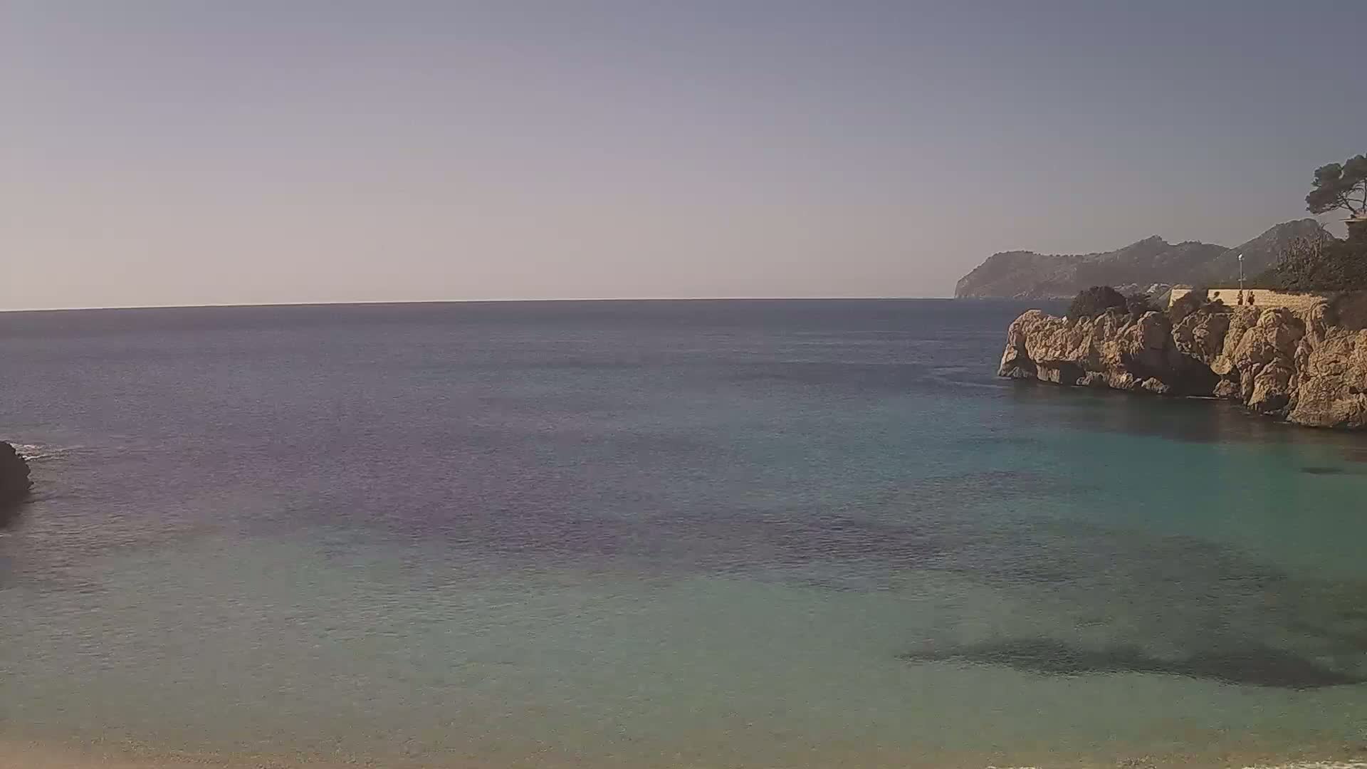 A tranquil cove with clear, shallow turquoise water, bordered by rocky cliffs and a distant mountain range under a clear sky.