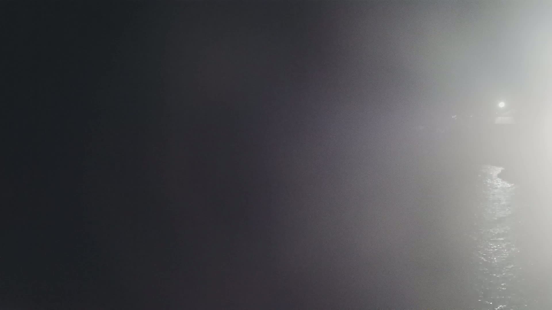 A dimly lit body of water is shrouded in dense fog at night, with a faint light visible in the distance.