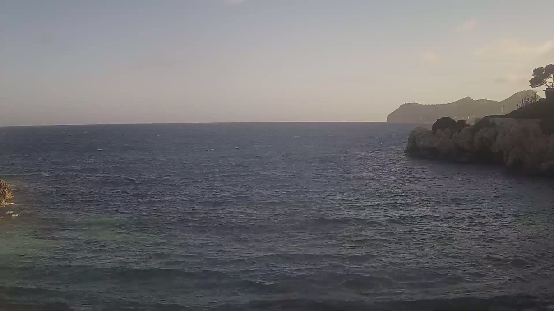 A calm, dark-blue sea stretches to a hazy horizon under a clear, light-grey sky, with rocky shores and distant hills visible on the right.