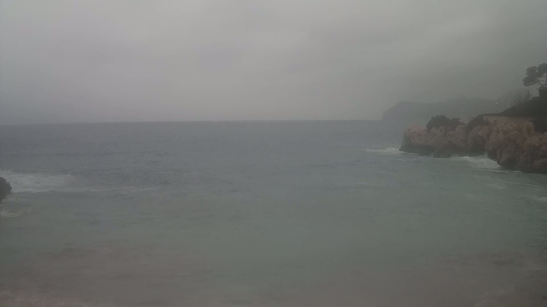 A hazy, overcast day shows a rocky shoreline with small waves lapping against it and a calm sea extending to a distant, indistinct landmass.