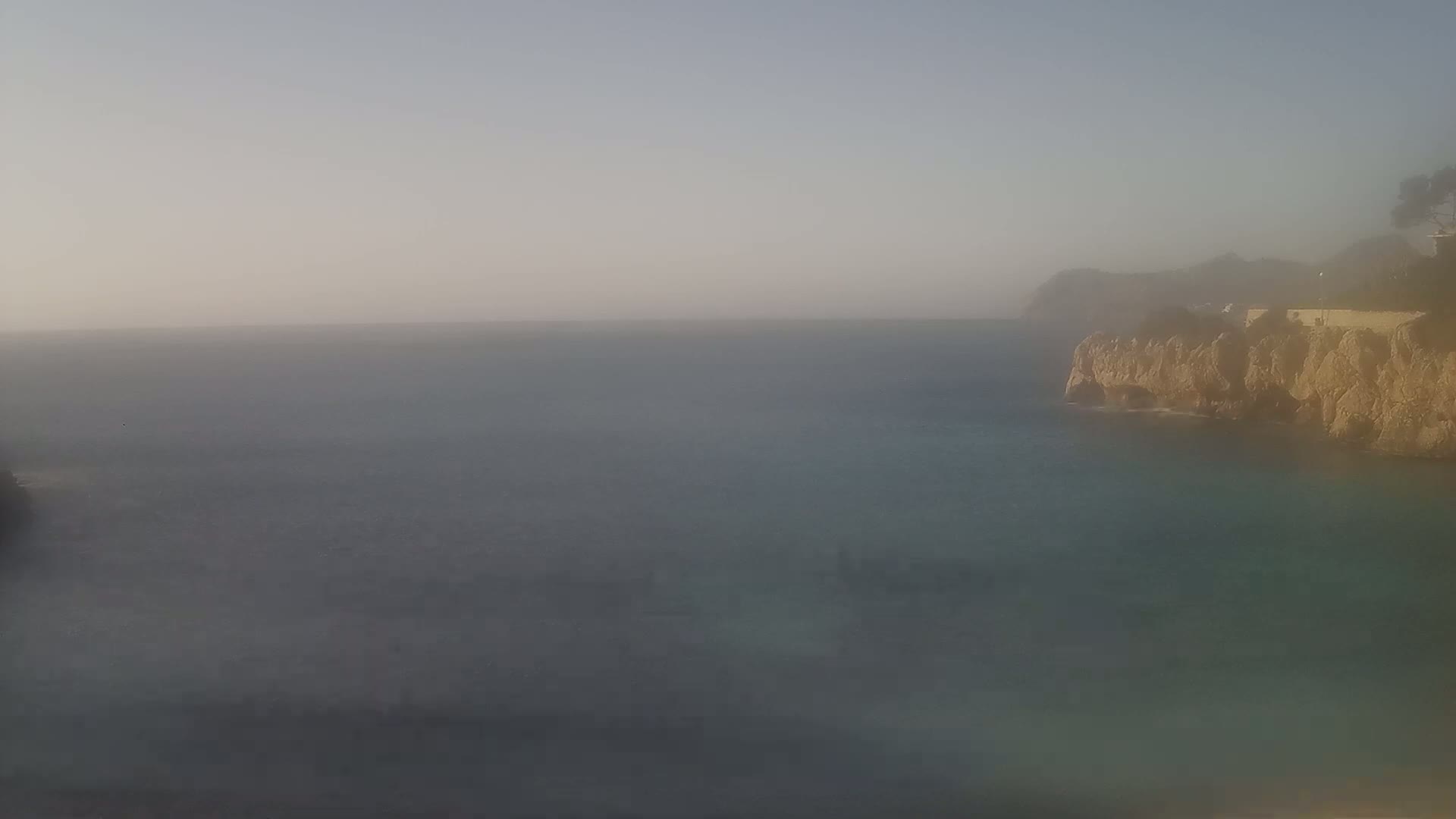 A hazy view of a calm sea meeting a rocky coastline under a light, overcast sky.