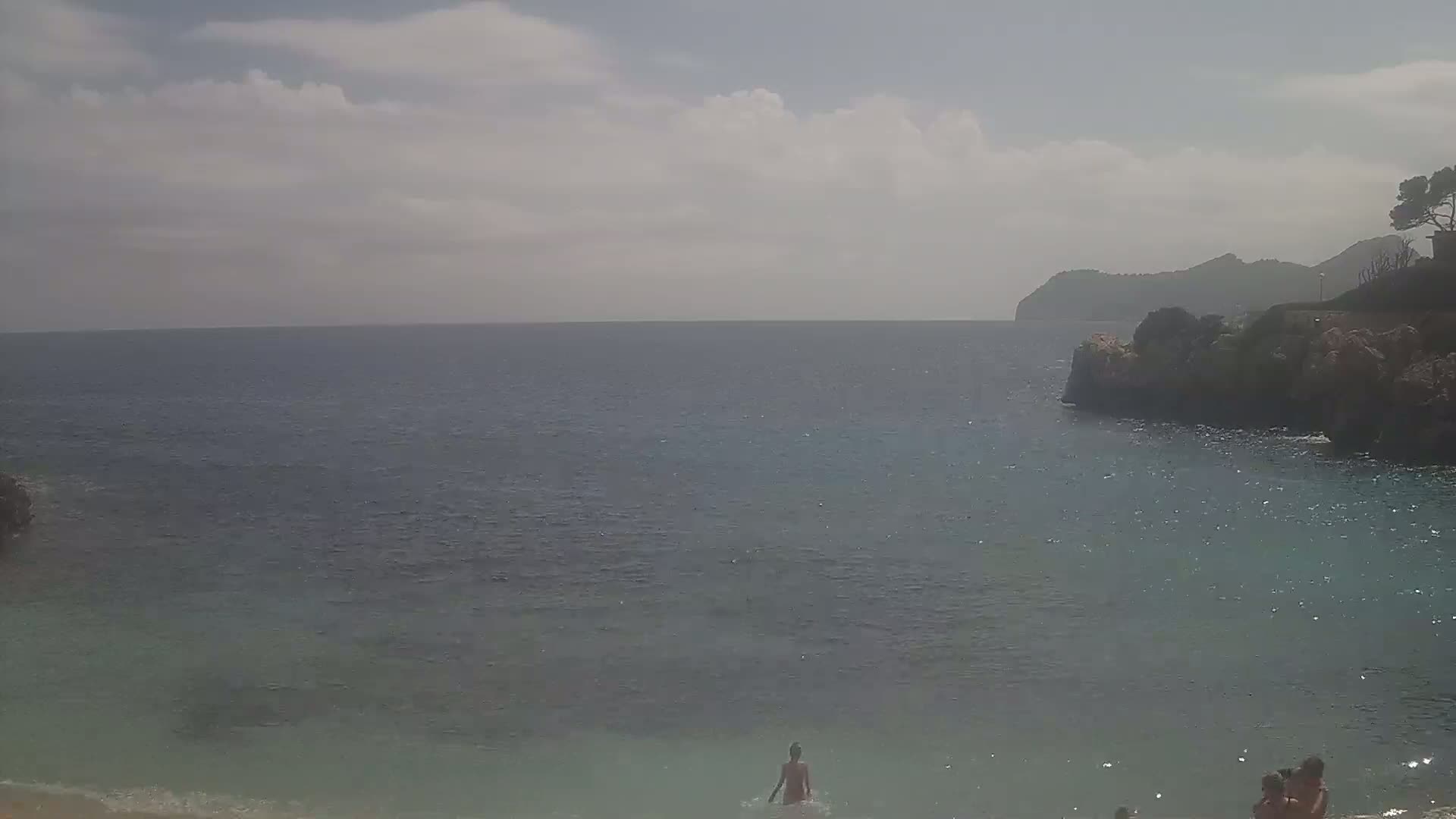 A partly cloudy day at a secluded beach cove with calm, clear water, where a few people are wading in the shallows and others are sitting near the shore.