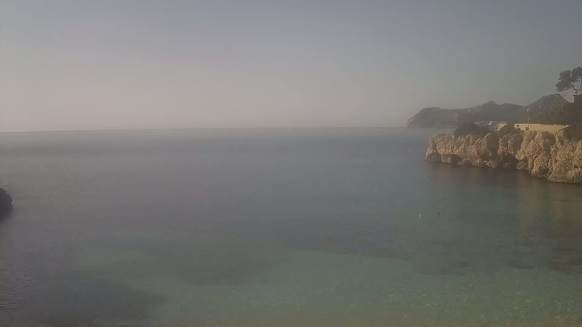 A hazy view of calm, clear water is partially enclosed by rocky cliffs, with a distant hillside visible under a light, hazy sky.