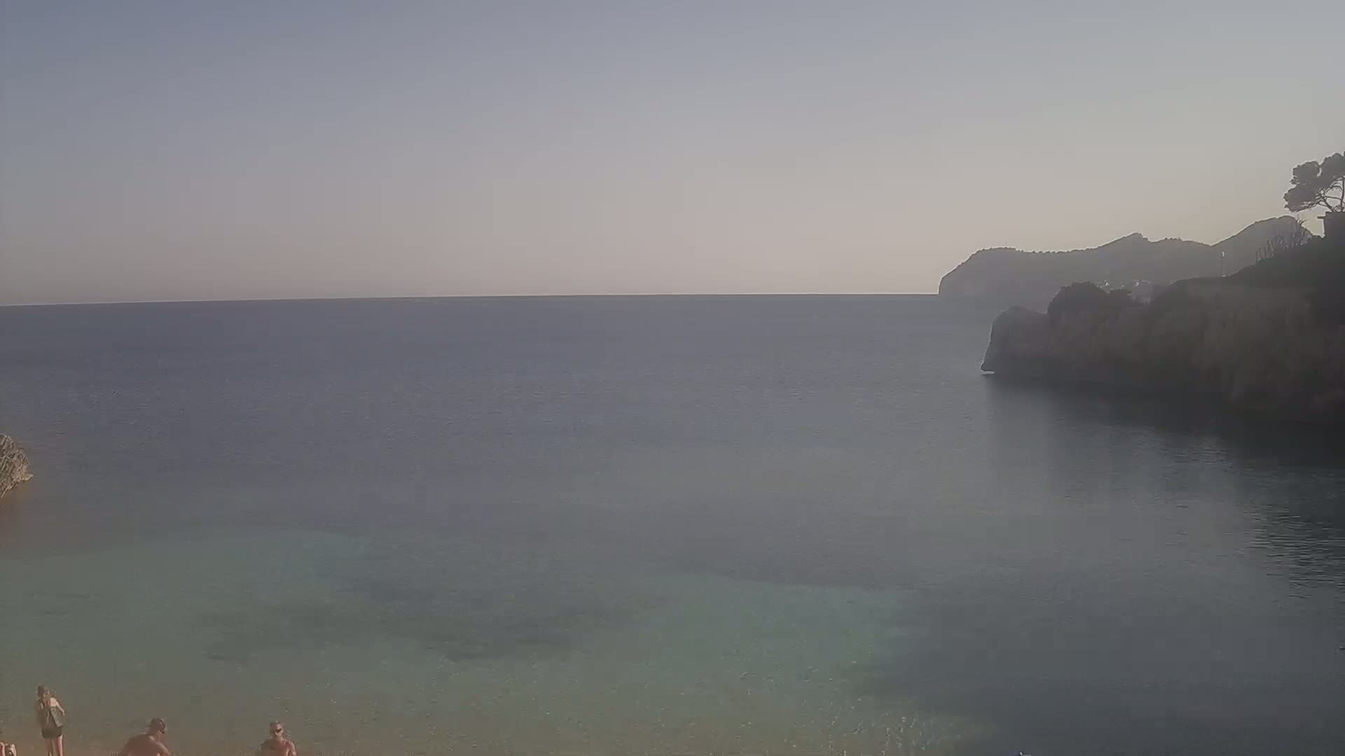 A calm, hazy day at a secluded cove with a few people on a sandy beach, overlooking a tranquil sea and distant hills.