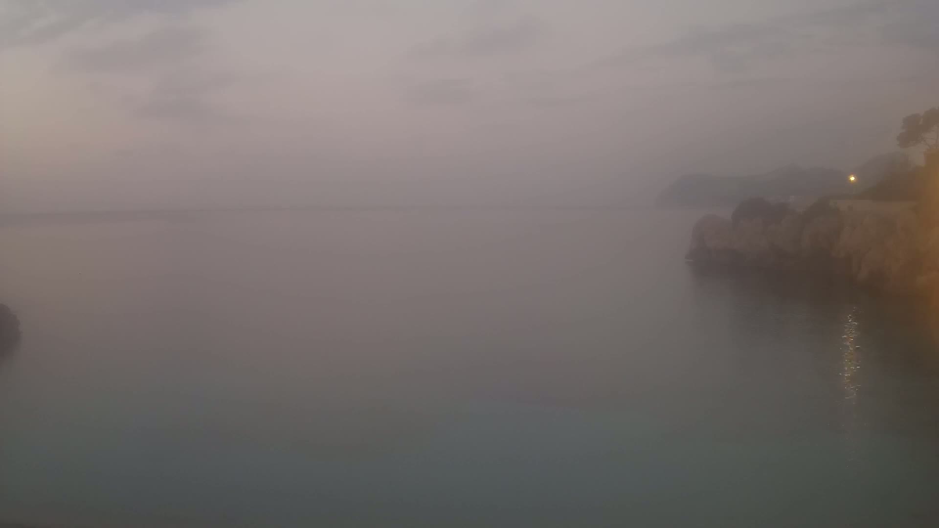 A foggy coastal scene shows a calm body of water with a rocky shore partially visible in the hazy conditions.