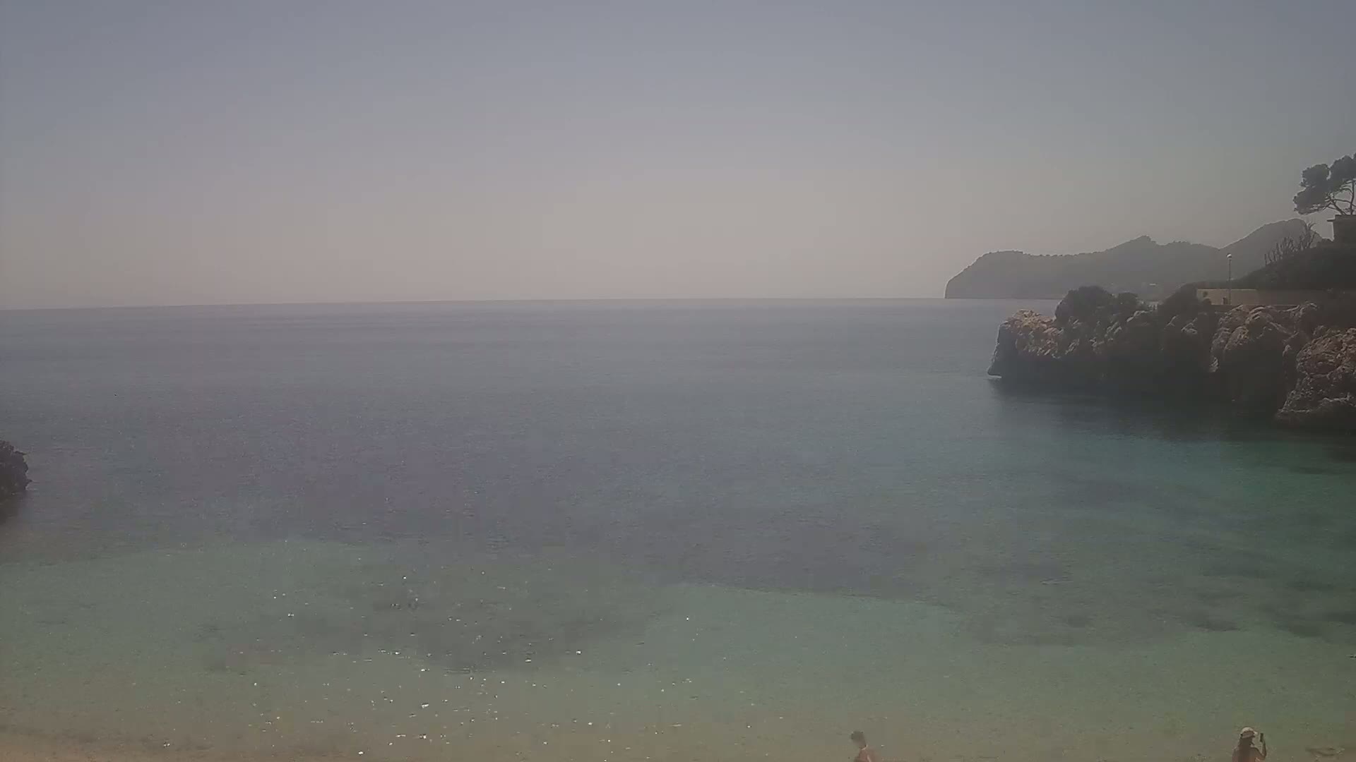 A hazy view of a calm, clear turquoise sea with rocky shores and a distant mountain under a light, bright sky.