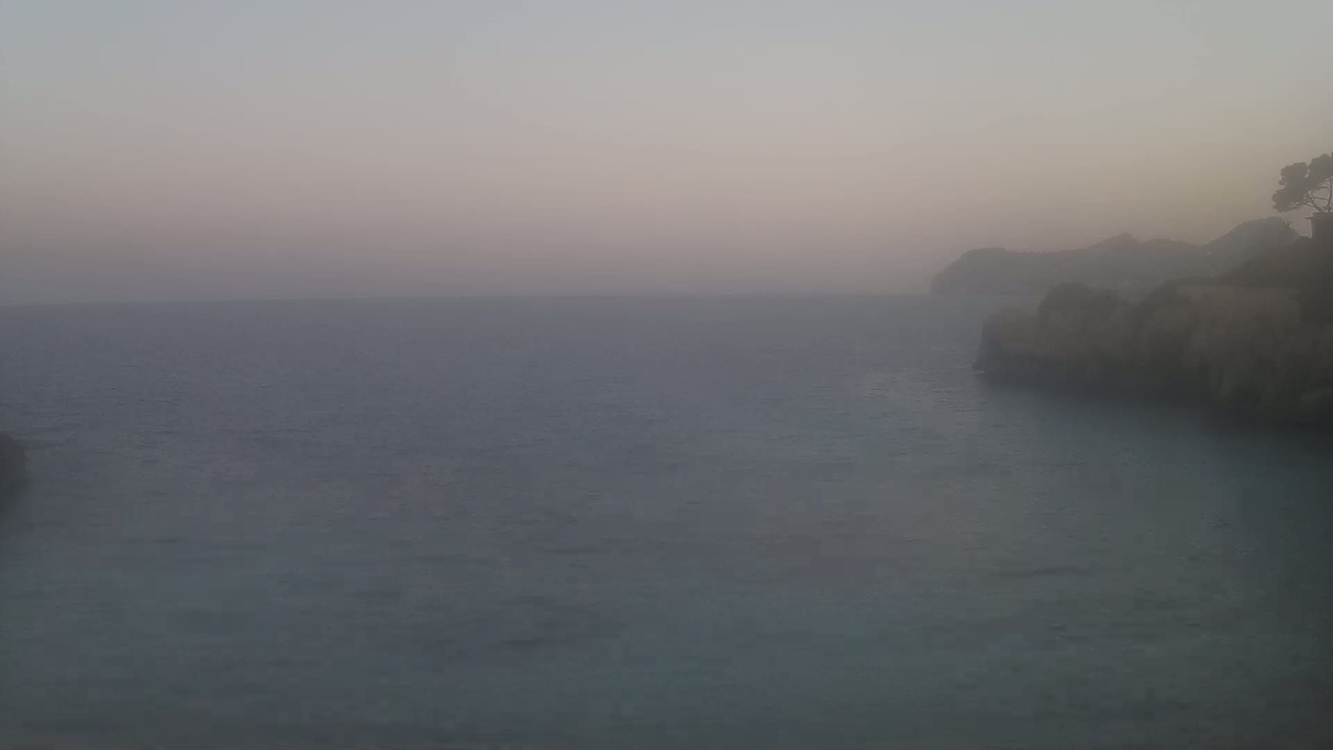 A hazy, calm sea stretches to a distant, indistinct coastline with a small, tree-topped cliff visible on the right.