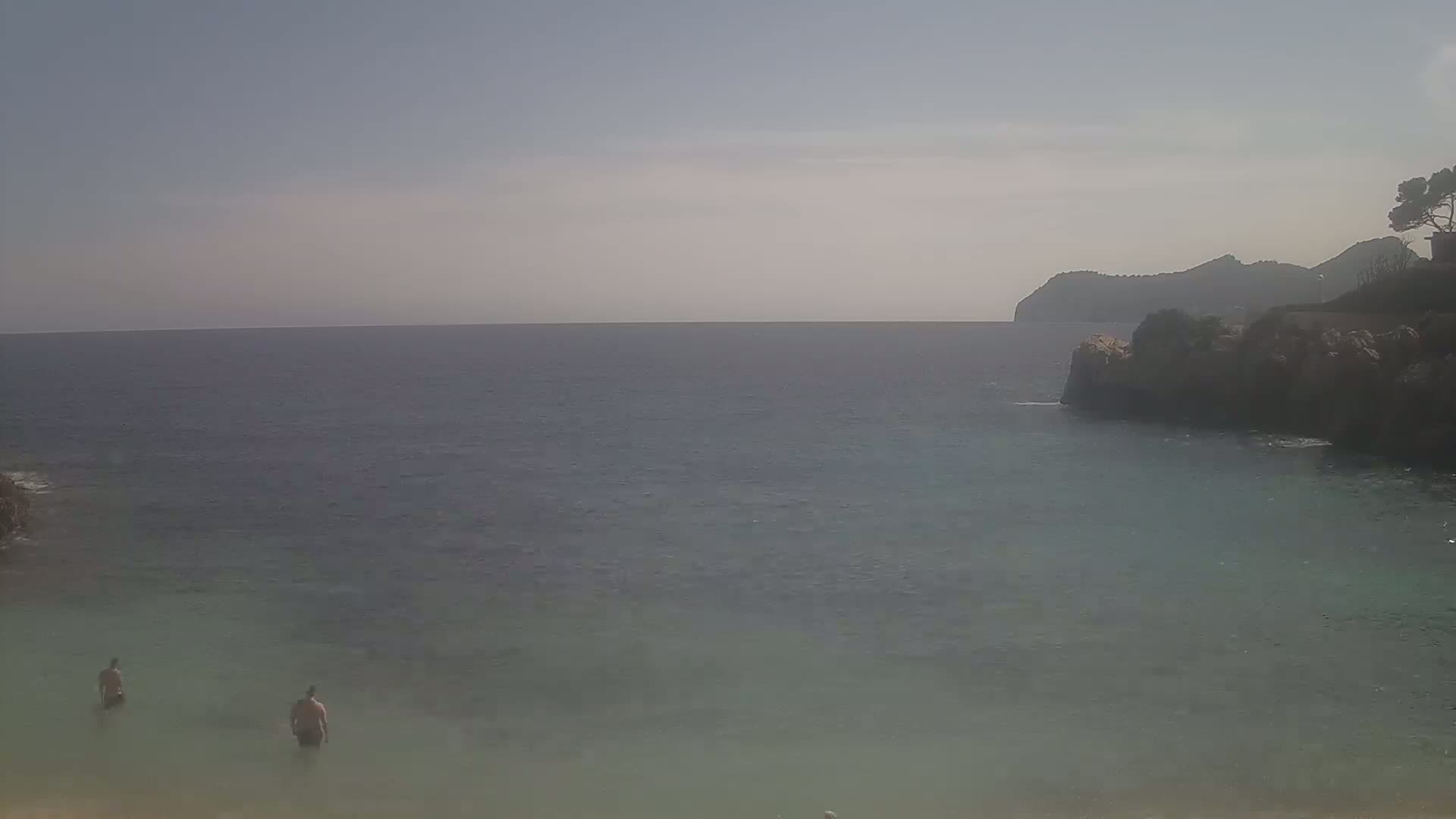 Two people wade in calm, clear water near a rocky shoreline, with a hazy distant mountain visible under a mostly clear sky.