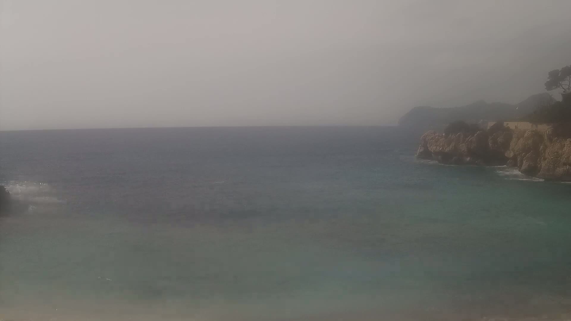 A hazy, overcast day reveals a rocky coastline meeting a calm, teal sea.