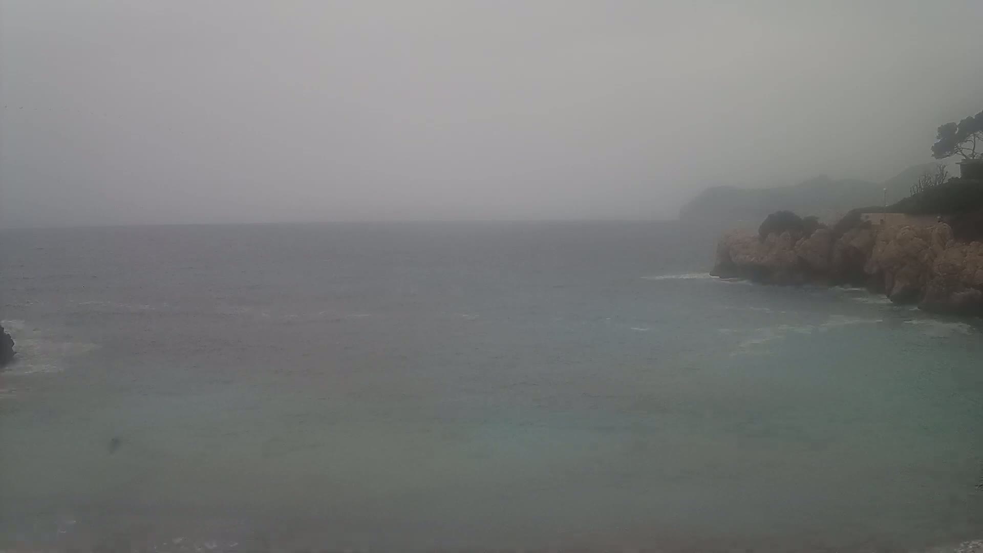 A hazy, overcast day shows a calm sea partially enclosed by rocky, tan-colored cliffs with sparse vegetation.