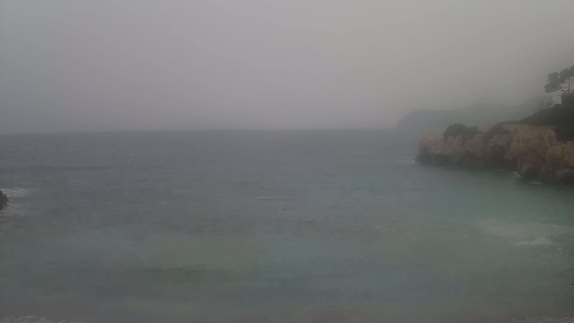 A hazy, grey day shows a calm sea meeting a rocky coastline with sparse vegetation.