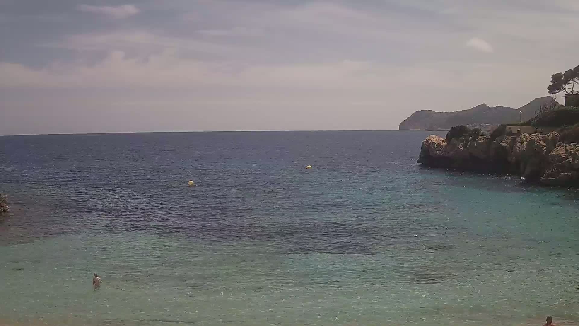 A calm, clear sea with a person wading in shallow water near a rocky shoreline and a distant mountain under a partly cloudy sky.