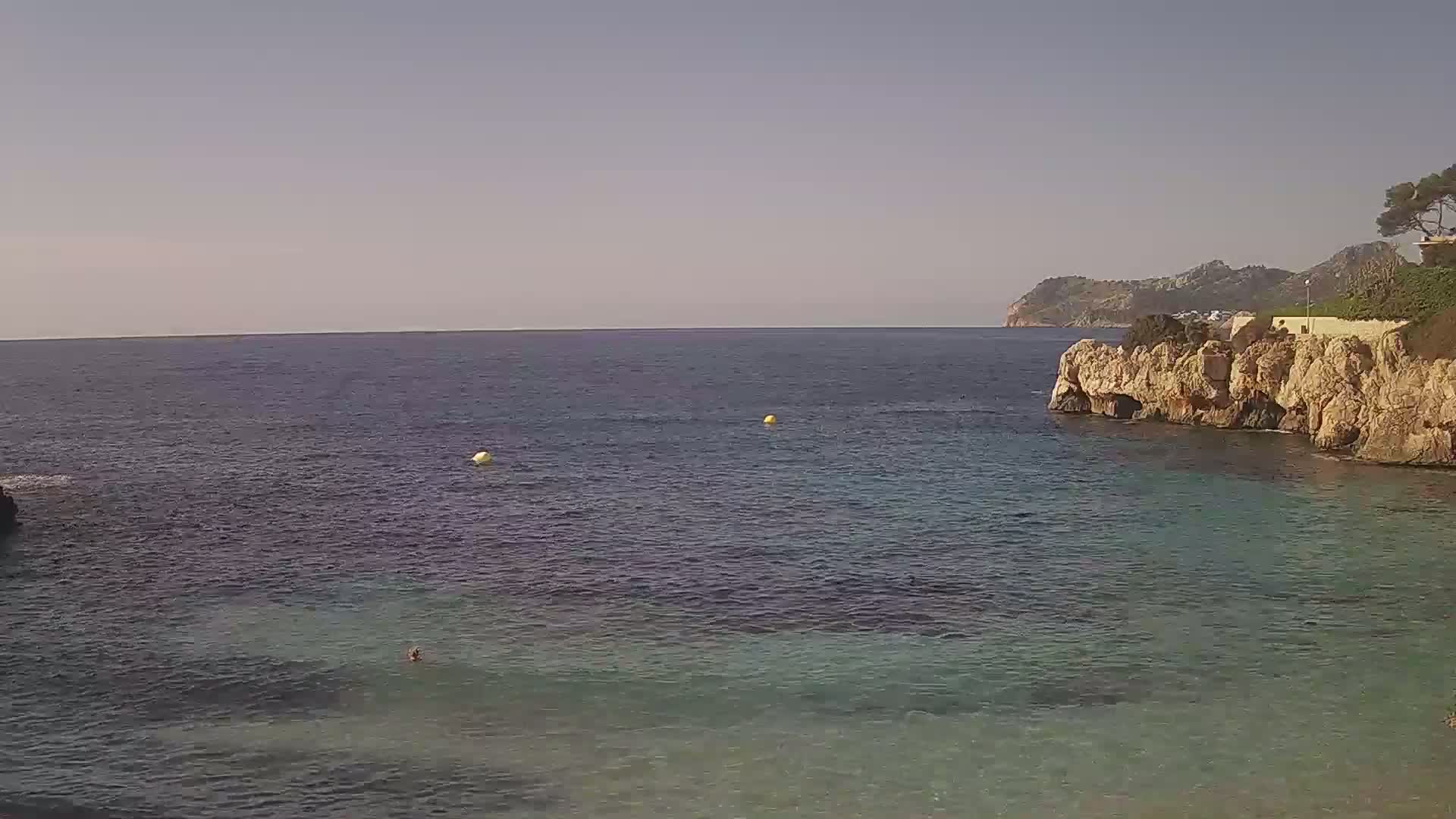 A calm, clear ocean cove is nestled between rocky cliffs on a sunny day, with a person swimming in the shallow water.