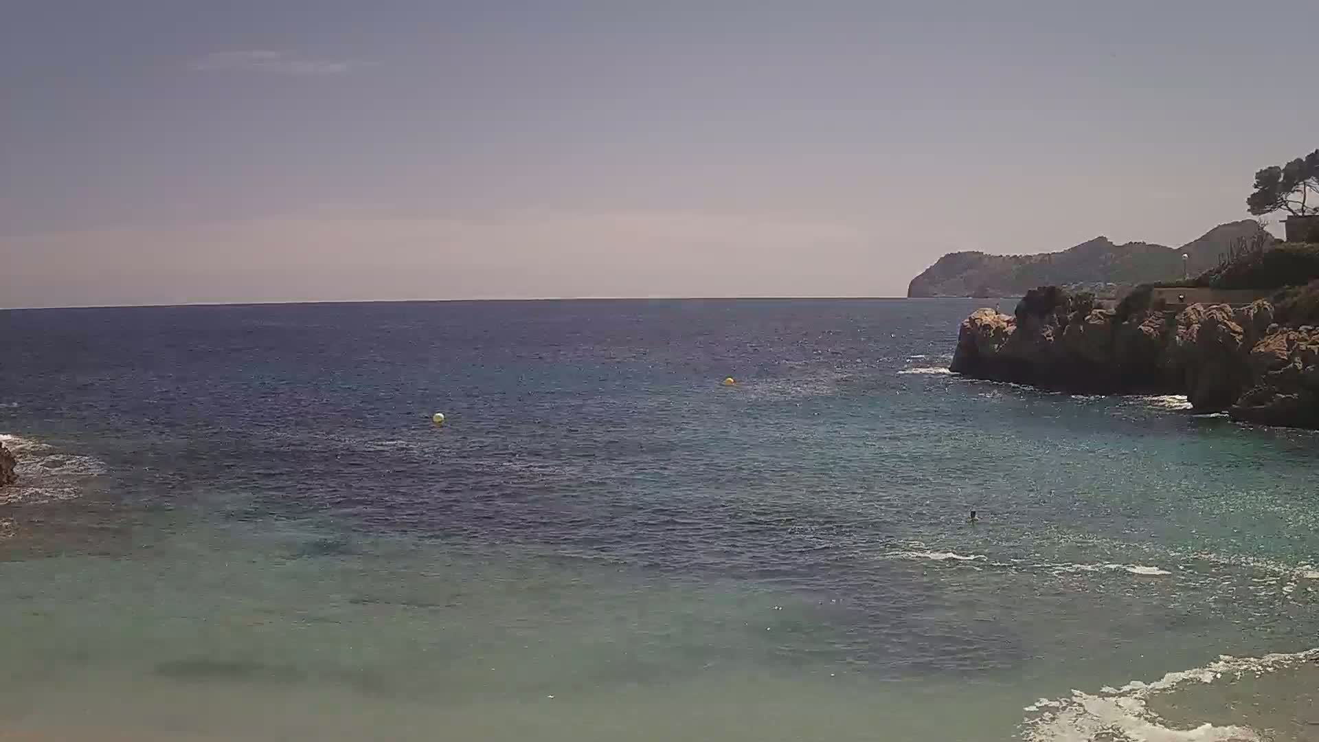 A calm, clear blue sea laps against rocky shores under a mostly sunny sky, with a person swimming in the bay.