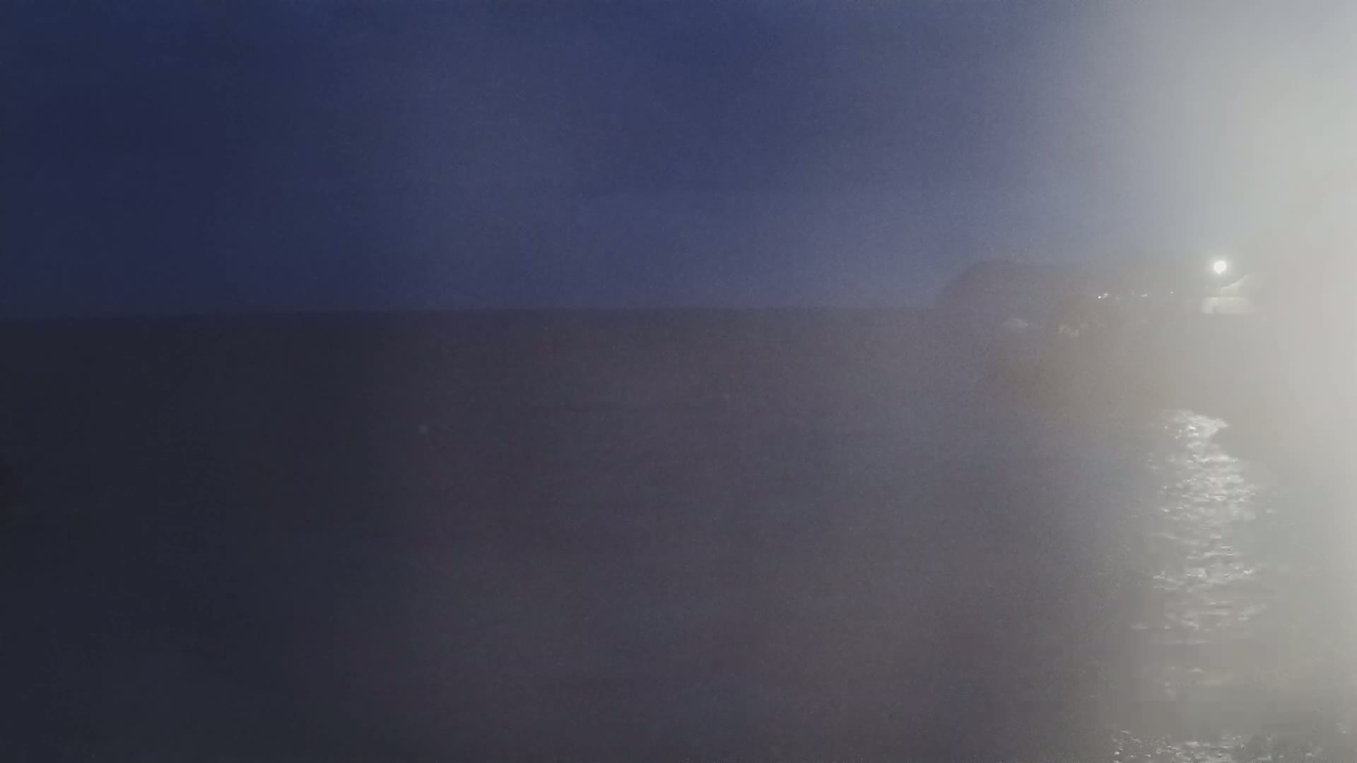 A dark, foggy scene shows a body of water at night with a dimly lit structure in the background.