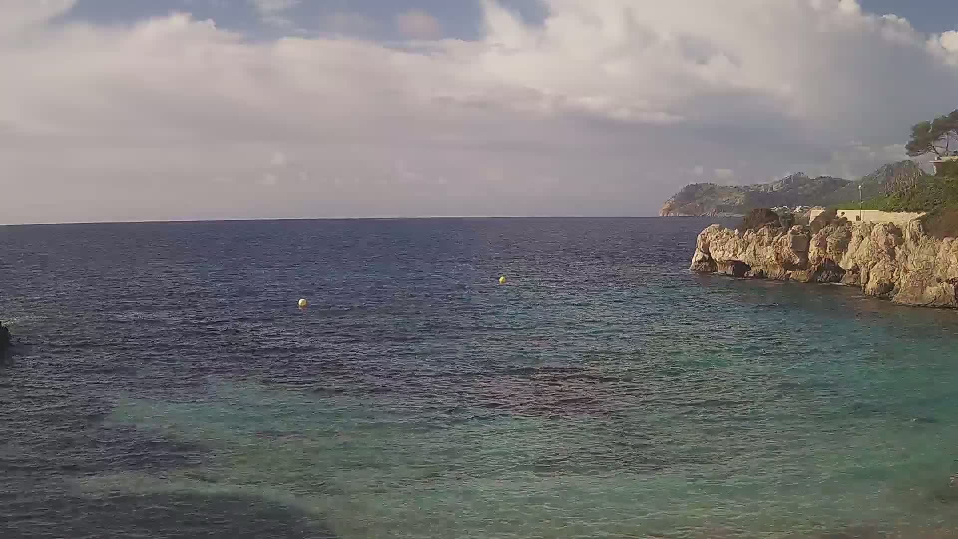 A partly cloudy sky overlooks a calm, teal sea partially enclosed by light-brown rocky cliffs and distant hills.