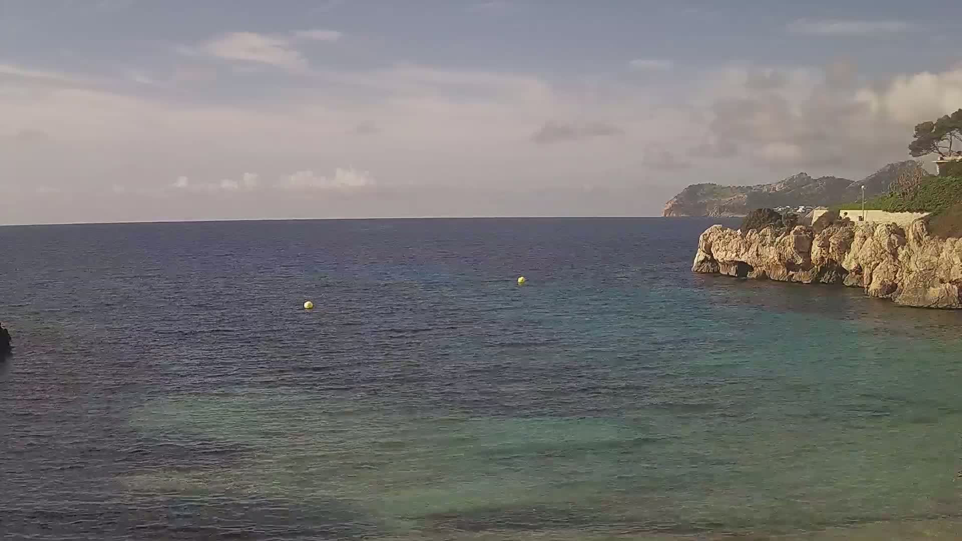 Calm, partly cloudy day shows a rocky cove with clear, teal water and a distant shoreline.