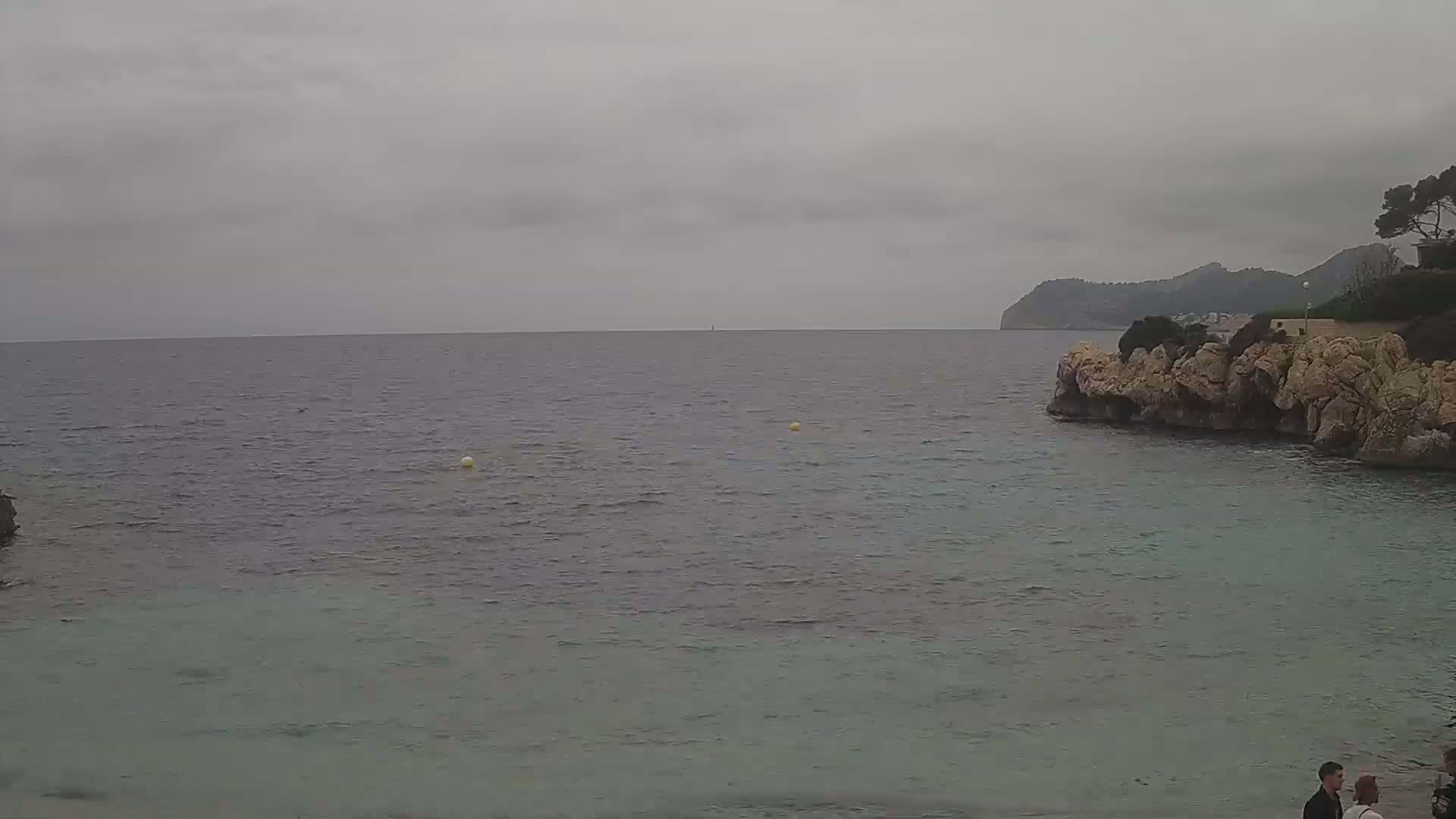 A calm, overcast day at a rocky cove with a few people standing near the shore, and a distant mountain visible across the water.