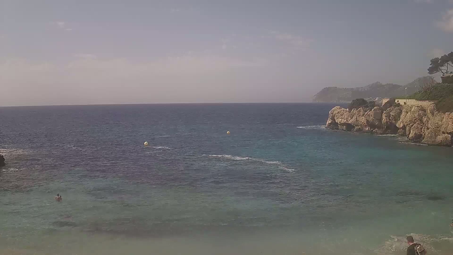 A calm, clear day shows a secluded cove with turquoise water, rocky cliffs, and a few people swimming or wading in the shallows.