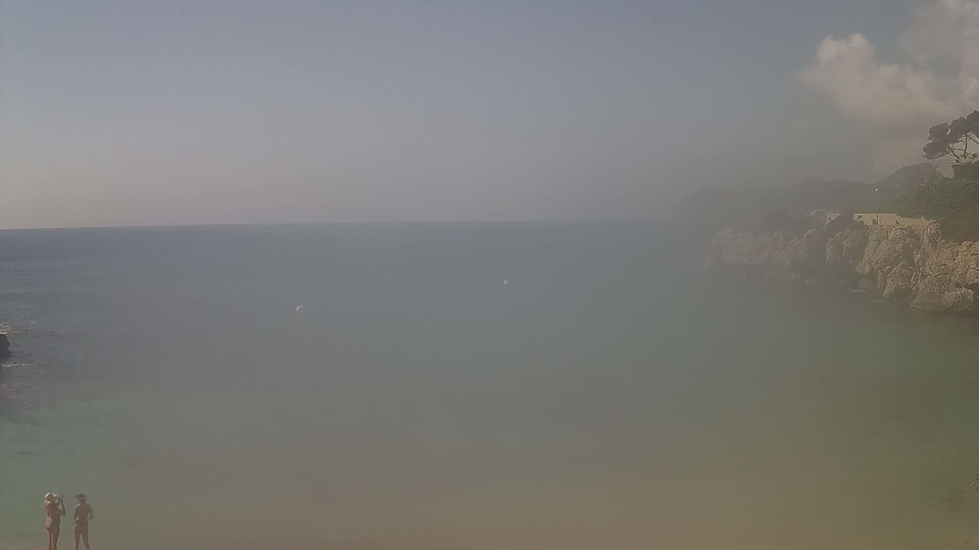 Two people stand on a hazy beach overlooking a calm, hazy sea partially enclosed by rocky cliffs and sparse vegetation.