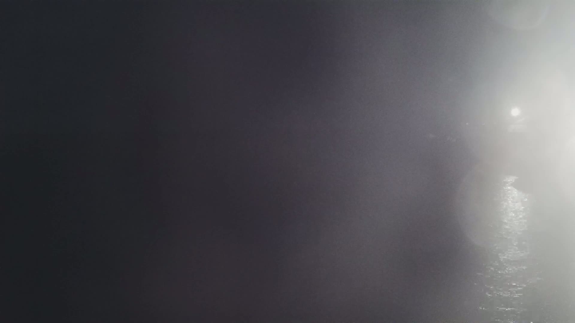 A dimly lit, foggy scene shows a dark body of water reflecting a light source in the distance.