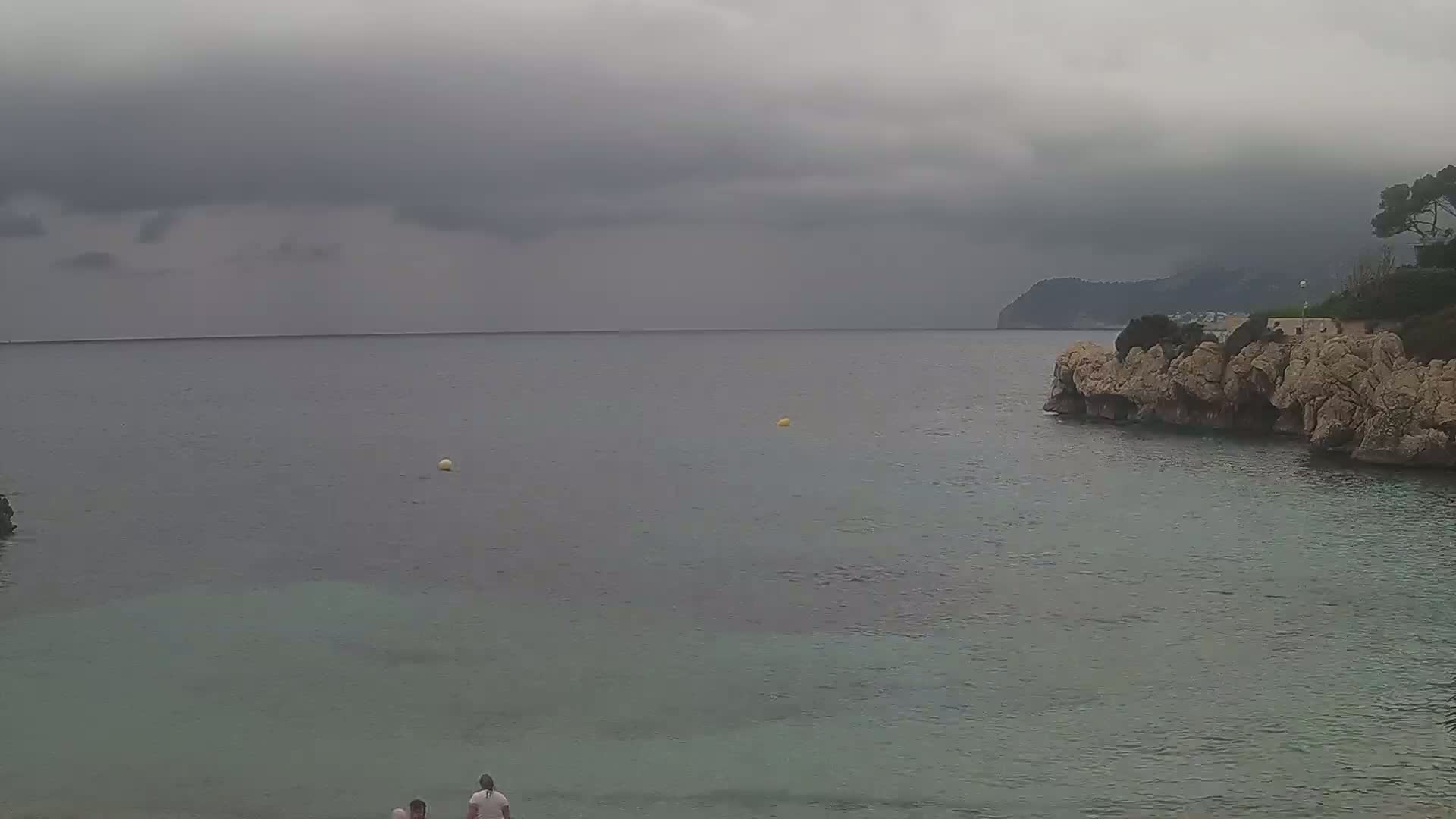 Two people stand on a rocky shore near calm, gray-green water under an overcast sky.