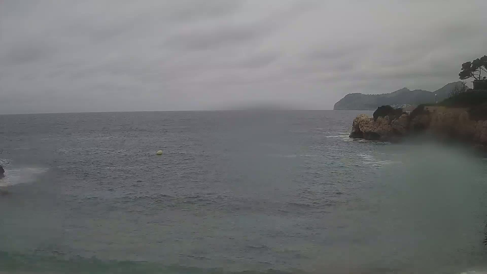 A calm sea under an overcast sky meets a rocky coastline with some vegetation on a hill in the background.