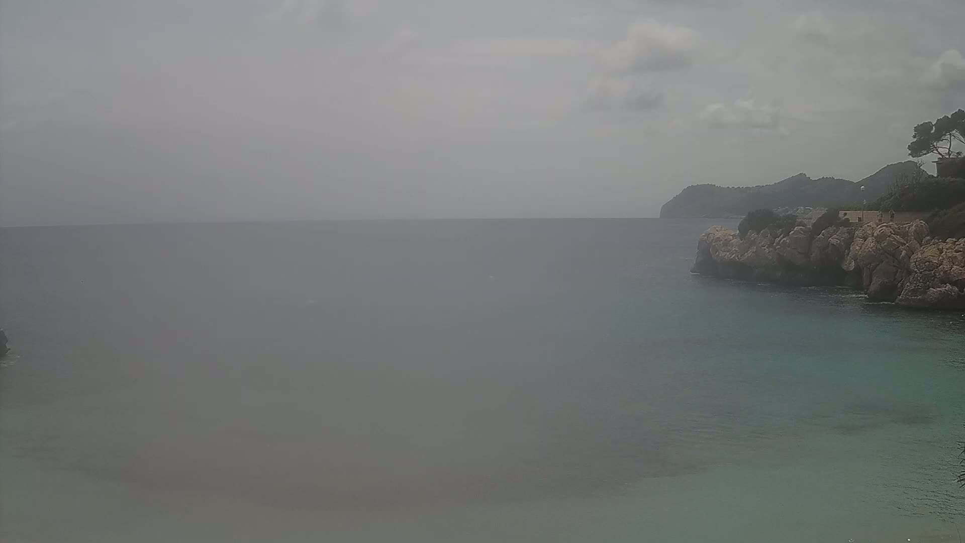 A hazy, overcast day reveals a rocky coastline meeting a calm sea, with a distant hill visible in the background.