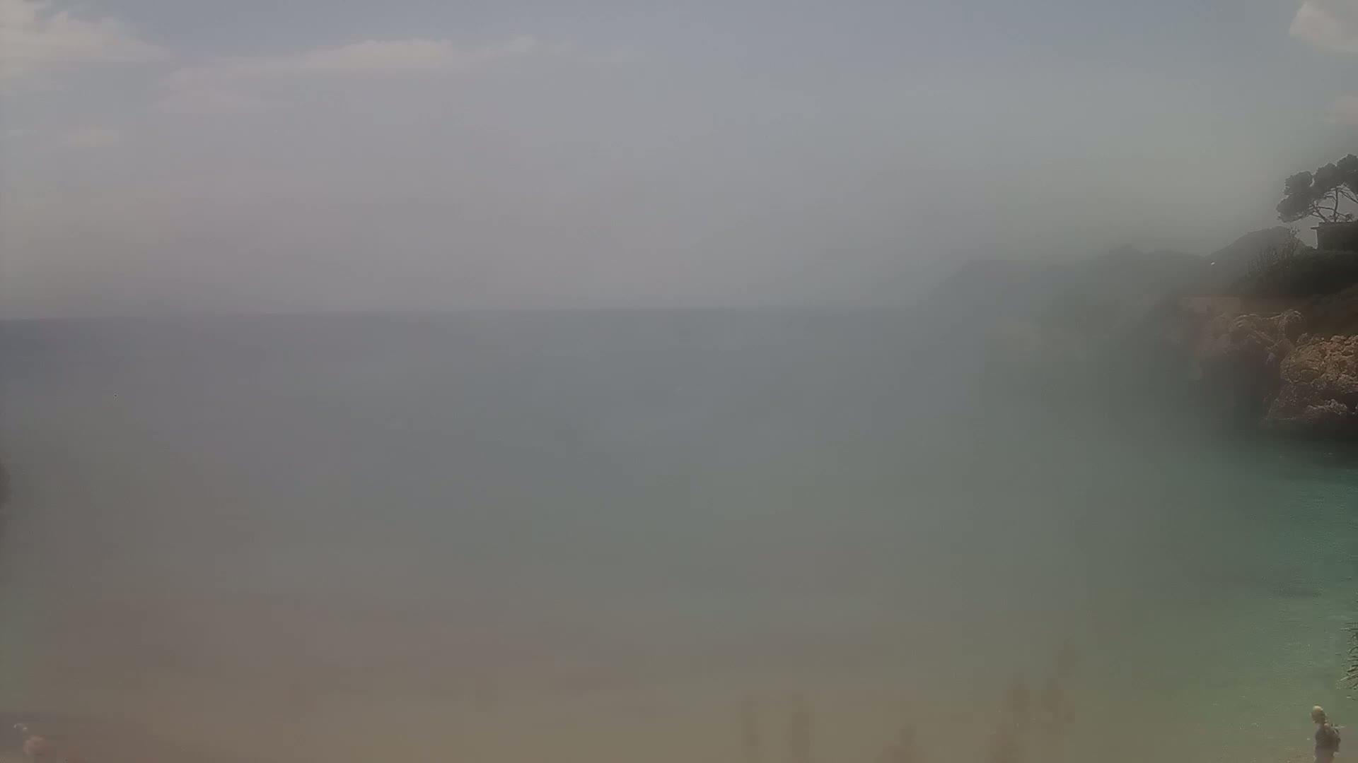 A hazy, overcast day at a beach cove shows a calm, somewhat obscured body of water partially surrounded by rocky cliffs with sparse vegetation.