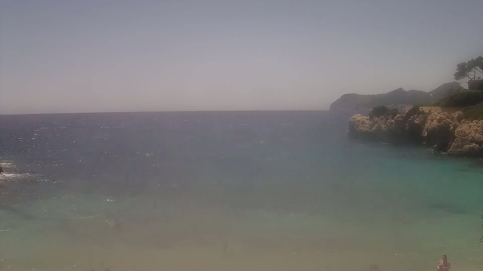 A mostly sunny day shows a calm, teal-colored cove with a rocky shore and a distant, hazy mountain range.