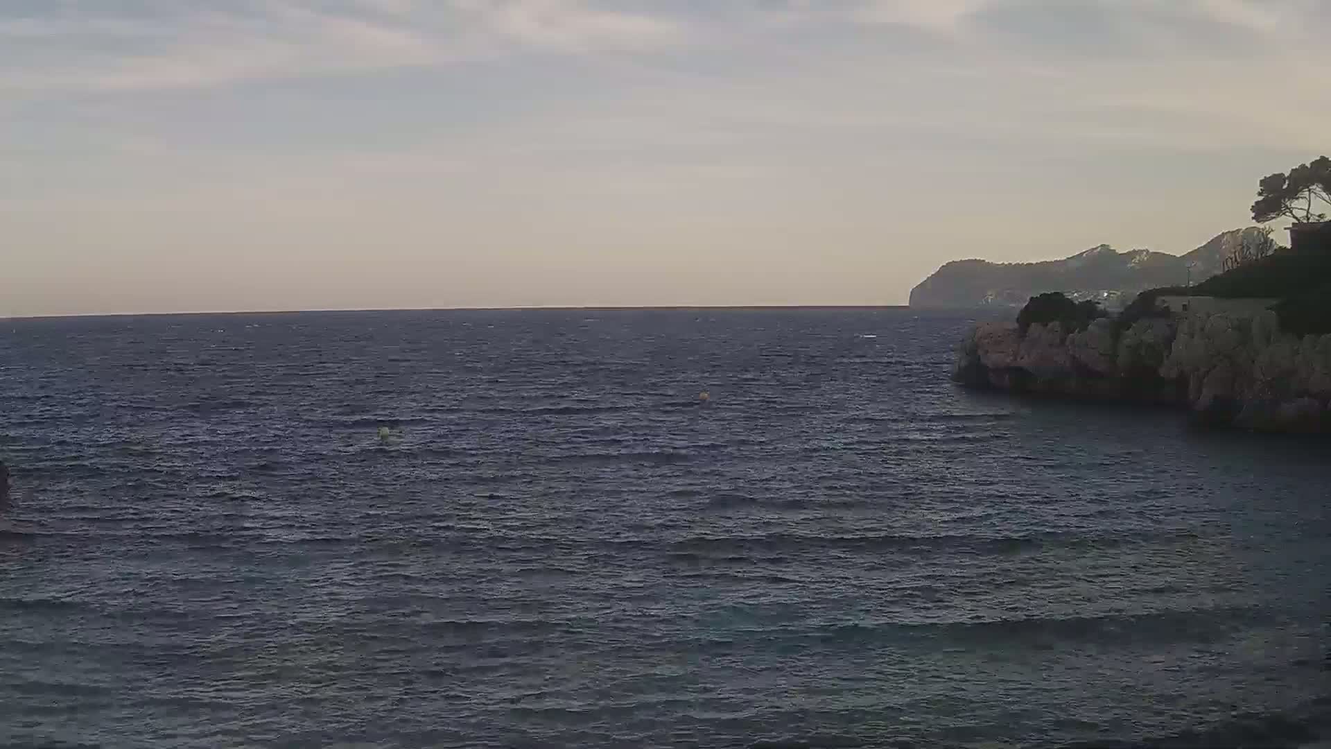 A partly cloudy sky overlooks a dark blue sea with small waves lapping against a rocky shoreline, where a small hill with some trees sits atop the cliffs.