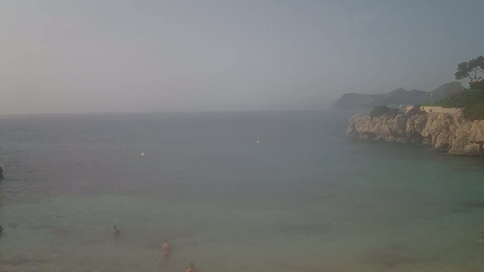 A hazy, calm sea laps against a rocky shore where a few people swim.