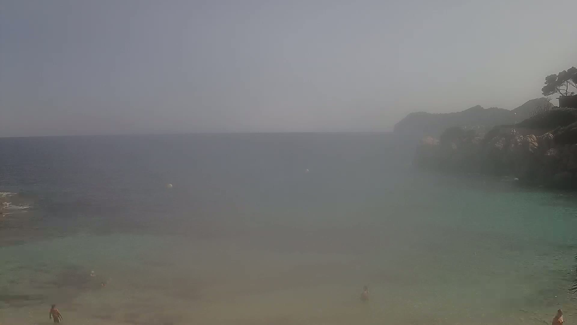 A hazy, calm ocean cove with a few people swimming and sunbathing near a rocky shore.