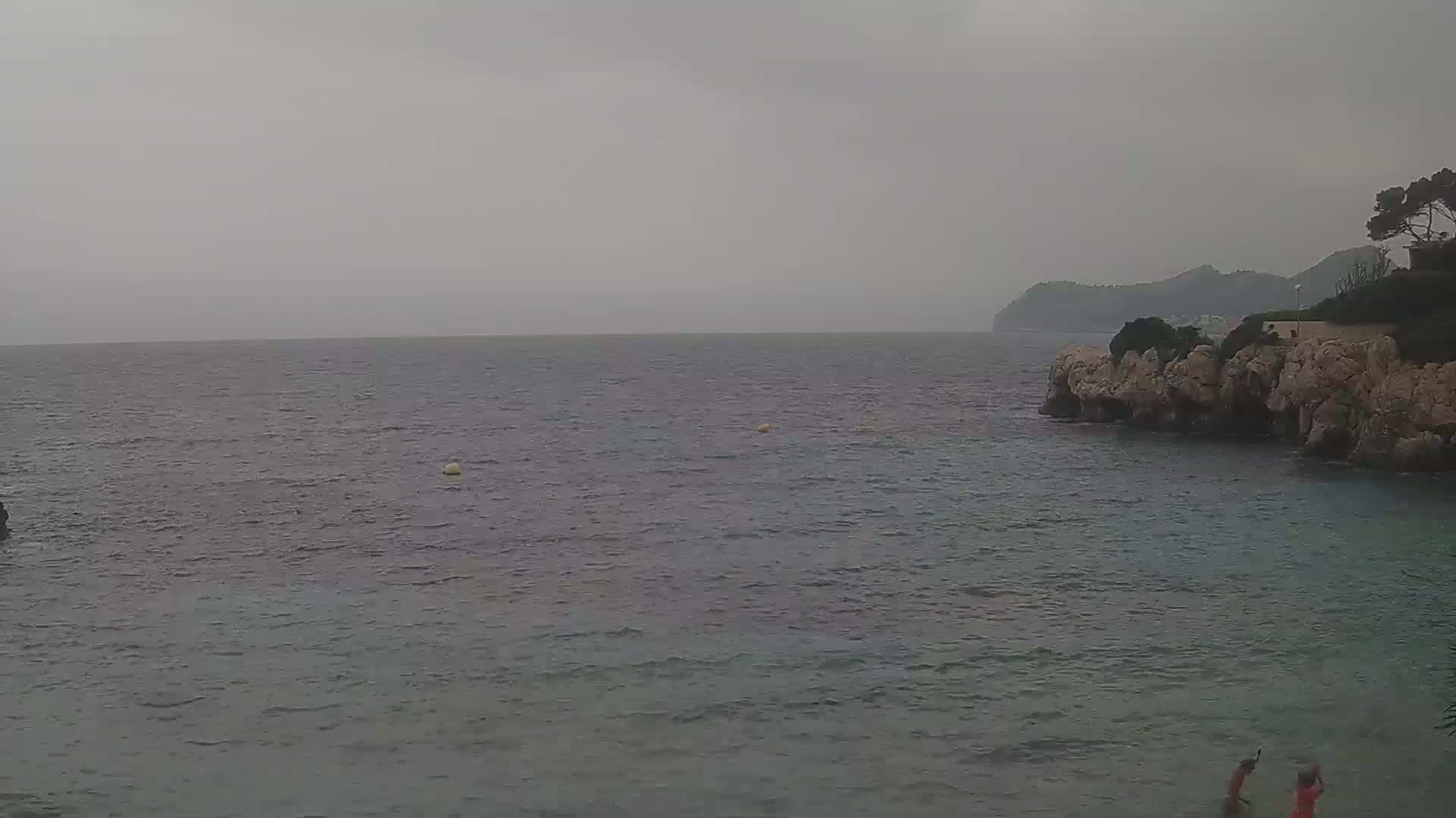 A calm, gray sea stretches to a distant, hazy coastline under an overcast sky, with rocky shores and a few people visible in the bottom right.