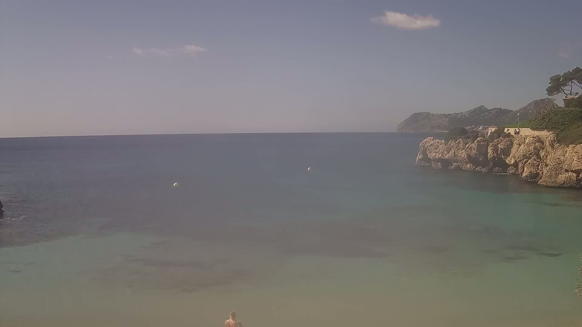 A calm, clear day at a secluded cove with light-colored rocky cliffs, a person standing on a sandy beach, and the sea extending to a distant, hazy horizon.