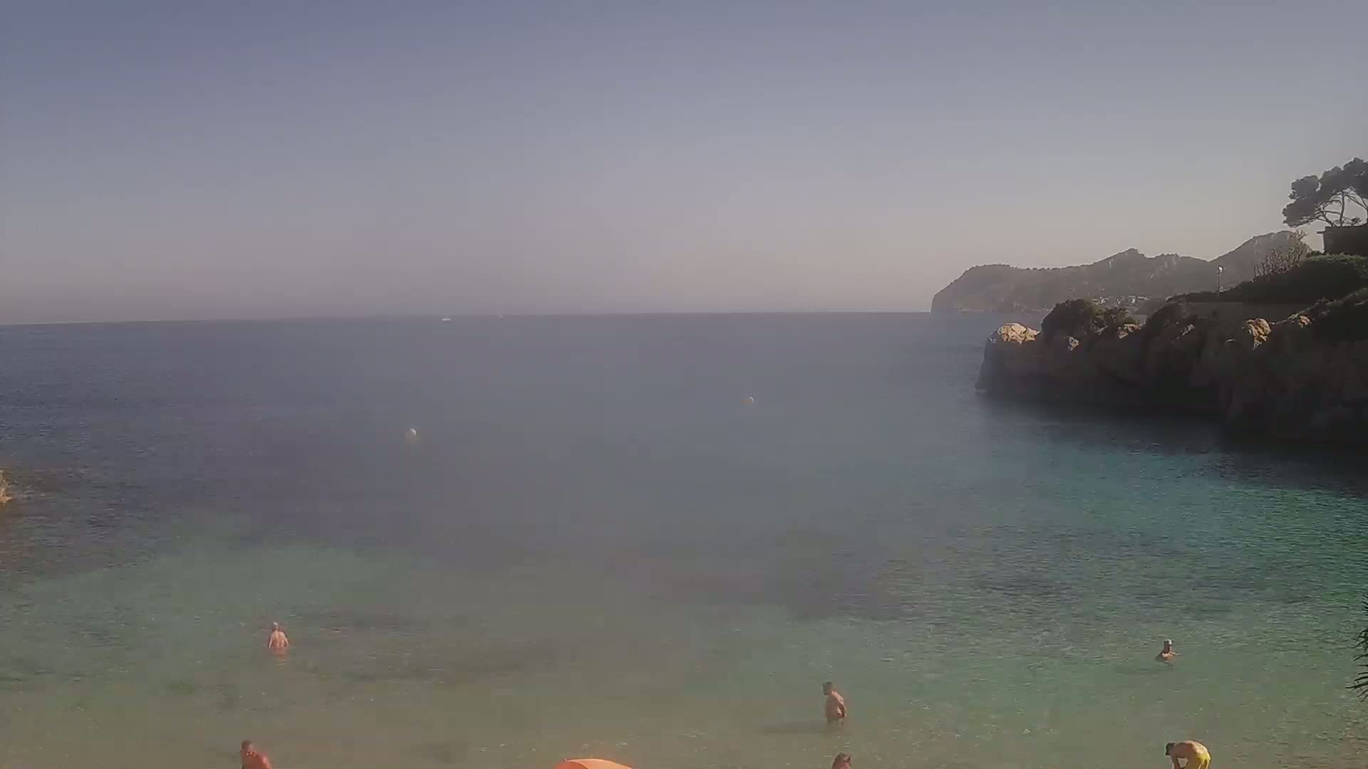 A hazy, sunny day at a secluded cove with calm, clear turquoise water, several people swimming and relaxing on the sandy shore, and a rocky cliff in the background.
