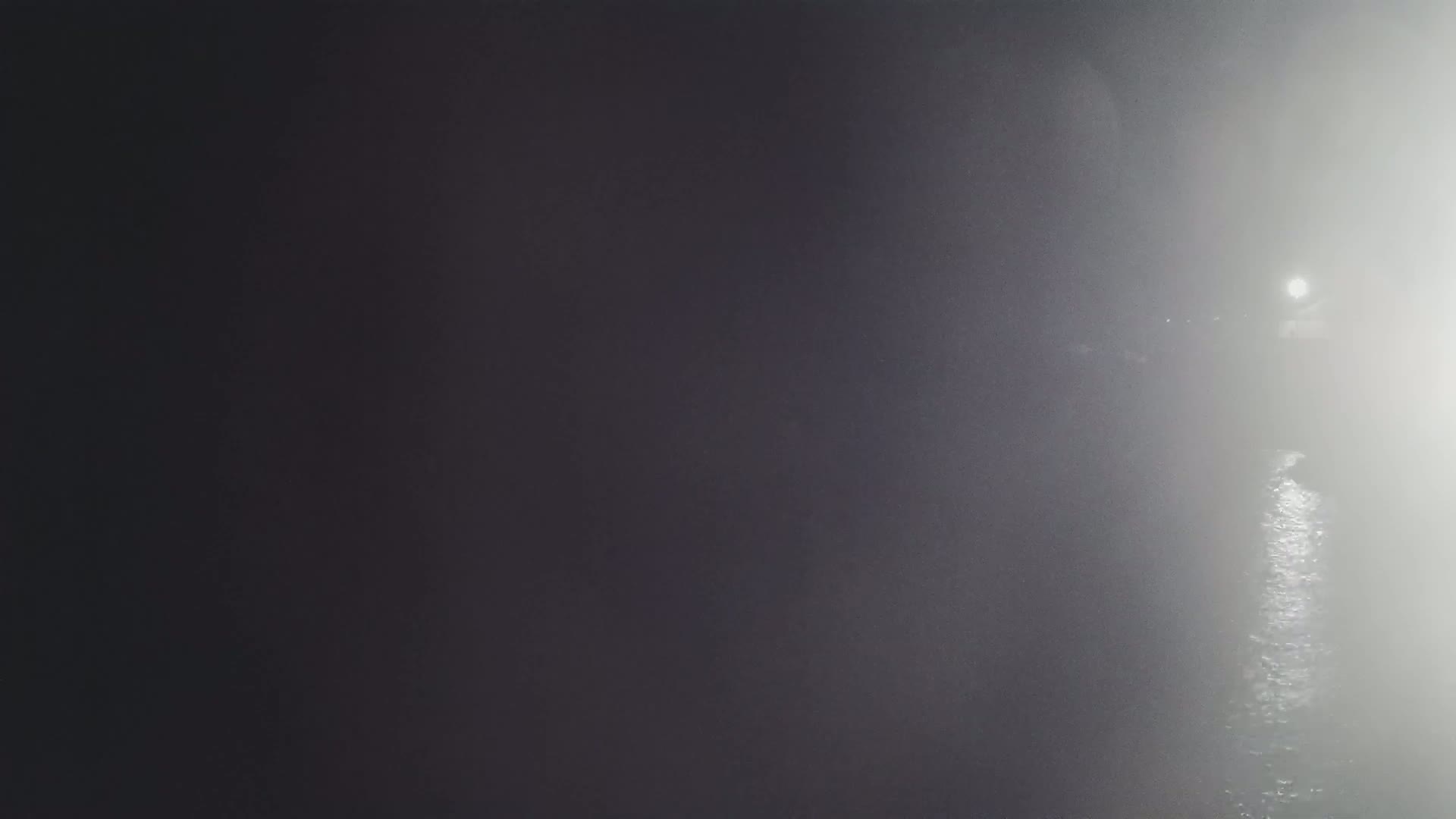A dimly lit, foggy scene shows a body of water reflecting a distant light source.