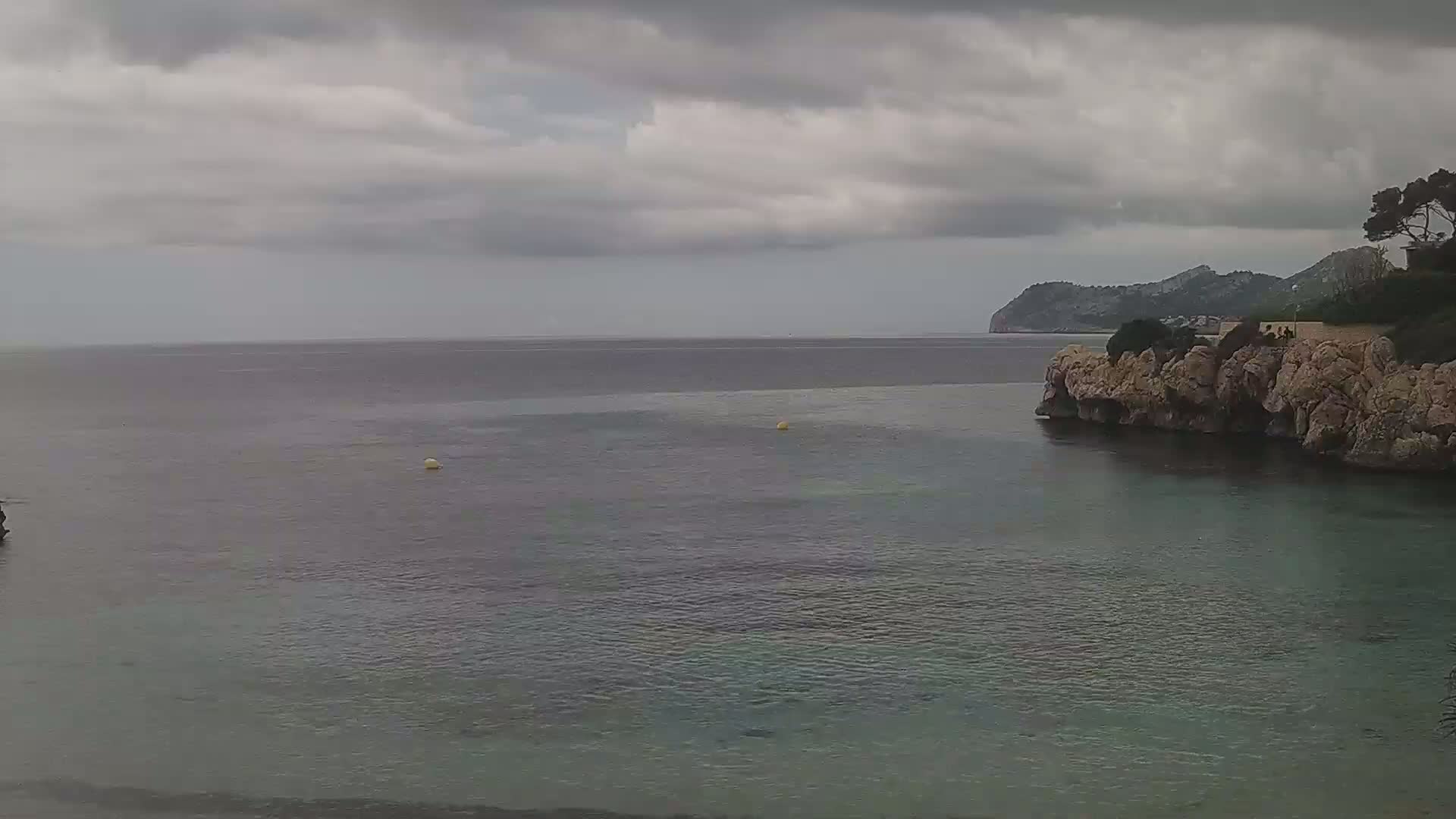 A calm sea under a cloudy sky is partially enclosed by a rocky shoreline with sparse vegetation.
