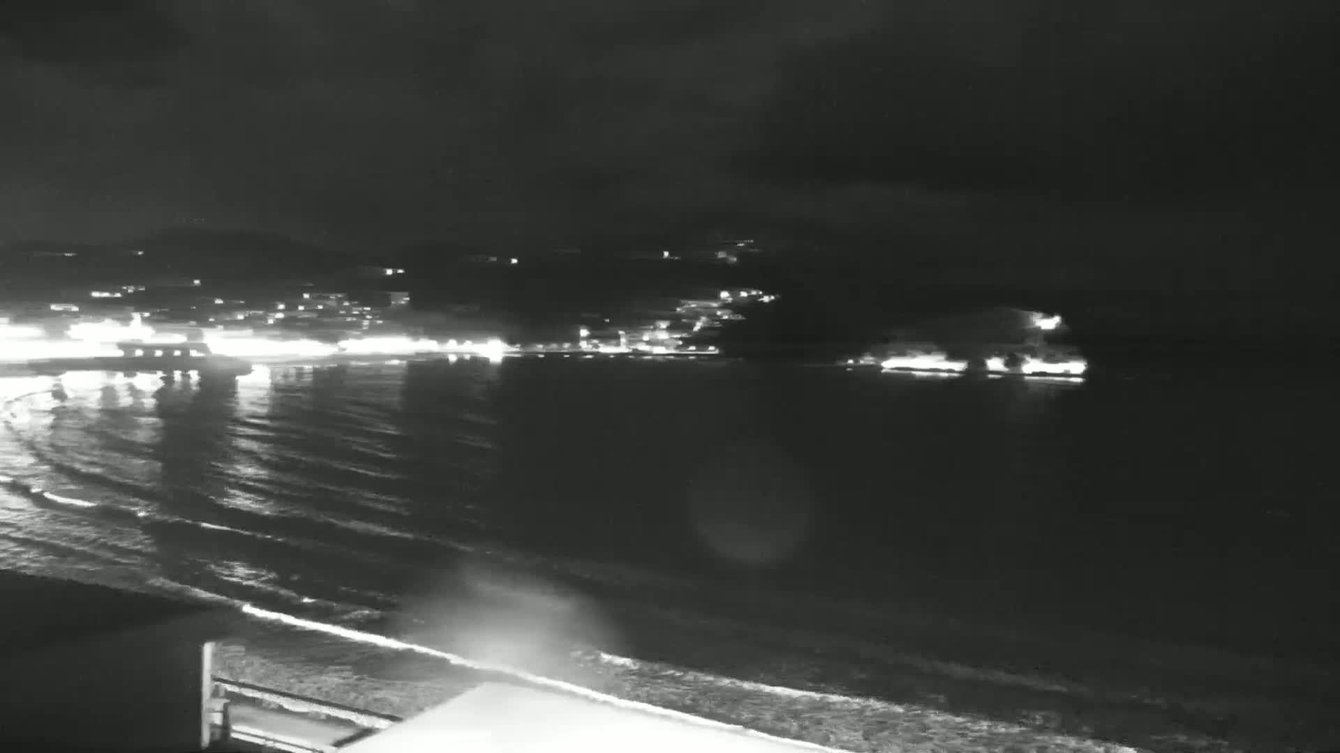 A nighttime view of a calm ocean with gentle waves lapping a shore, overlooking a distant town with lights illuminating the coastline under a dark sky.